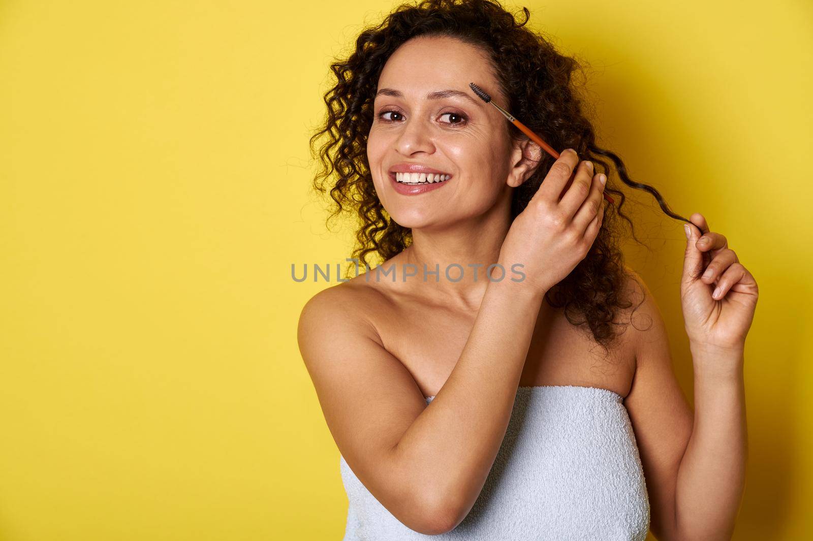 Smiling mixed race young woman with perfect skin and makeup posing with makeup brush over yellow background with space for text