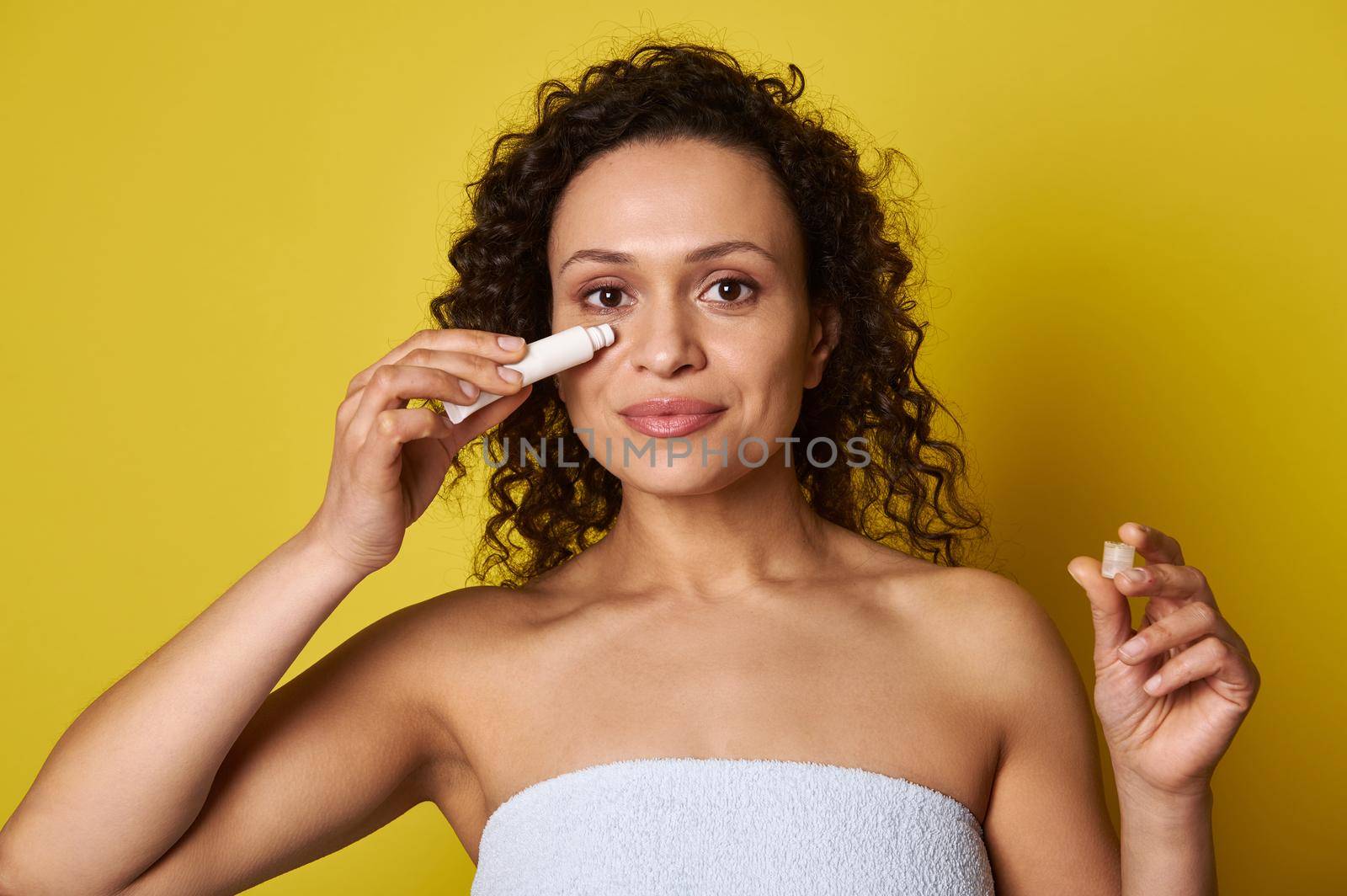 Close-up portrait of an attractive natural beauty woman wrapped in a white bath towel holding a tube of cosmetic product and applying it under the eyes by artgf