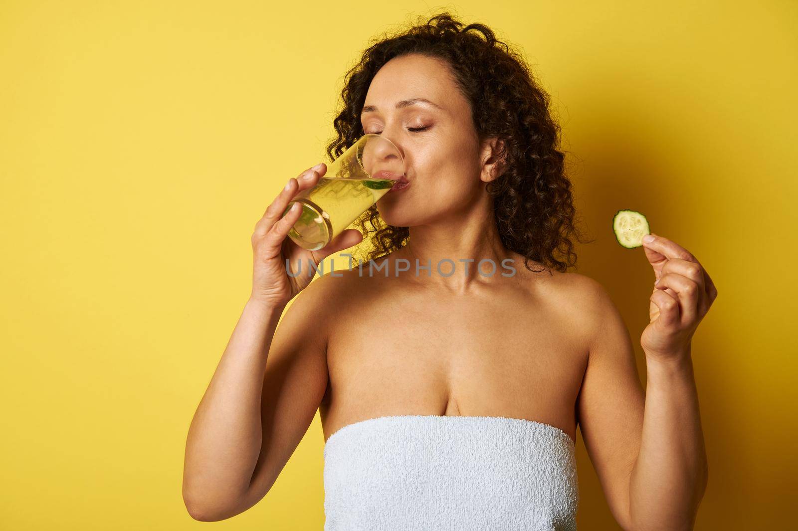 Young half-naked woman, wrapped in a towel, holding a slice of cucumber and drinking water from a glass, standing against a yellow background with space for text
