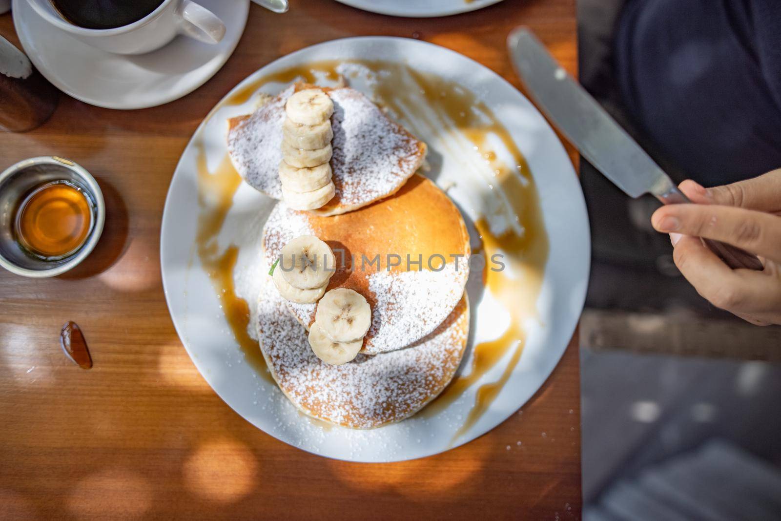 Top view of hand holding knife above tasty pancakes with powdered sugar, syrup, and banana slices. Sunlight on delicious sugary dessert with fruit on wooden table. Sweet breakfast and food
