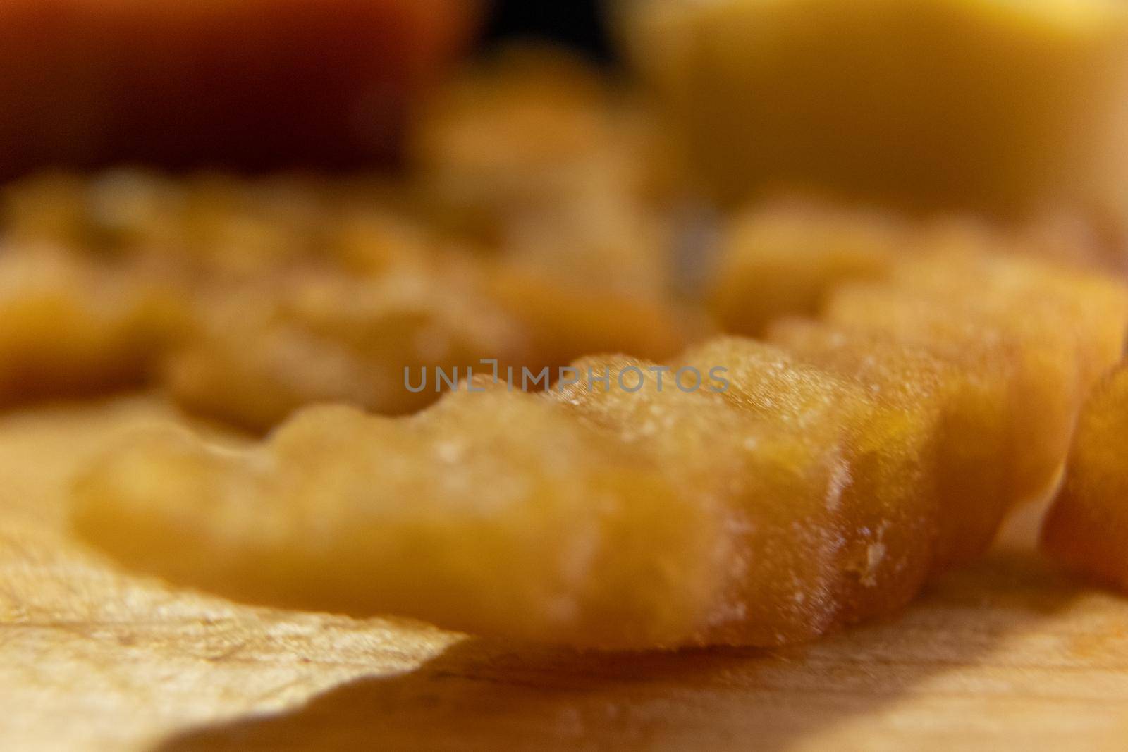 Extreme close-up of wavy French fry on wooden surface with blurry cups of ketchup and melted cheese as background. Tasty fried snacks and two glasses of red and yellow dressing. Delicious fast food