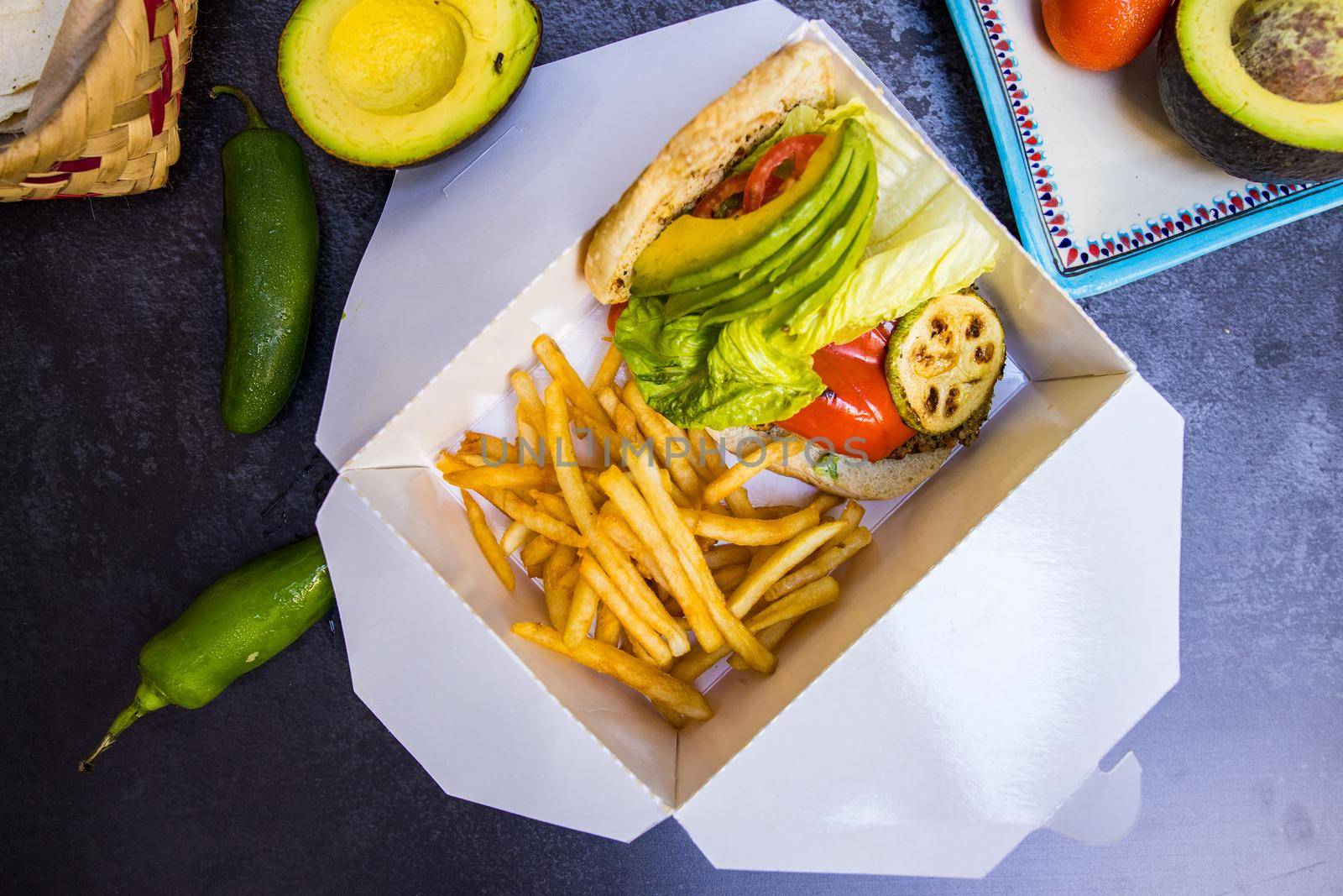 Top view of tasty French fries and sliced vegetables in white box above dark surface. Delicious avocado, tomato, lettuce, chili peppers, and fried snacks from above. Food and healthy meals