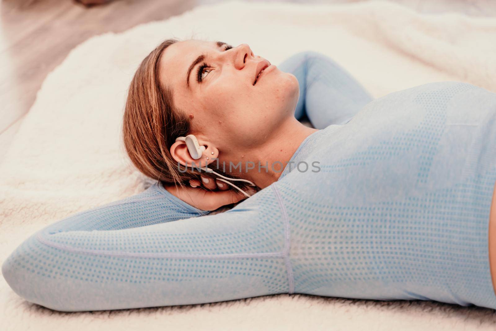 Side view portrait of relaxed woman listening to music with headphones lying on carpet at home. She is dressed in a blue tracksuit. by Matiunina