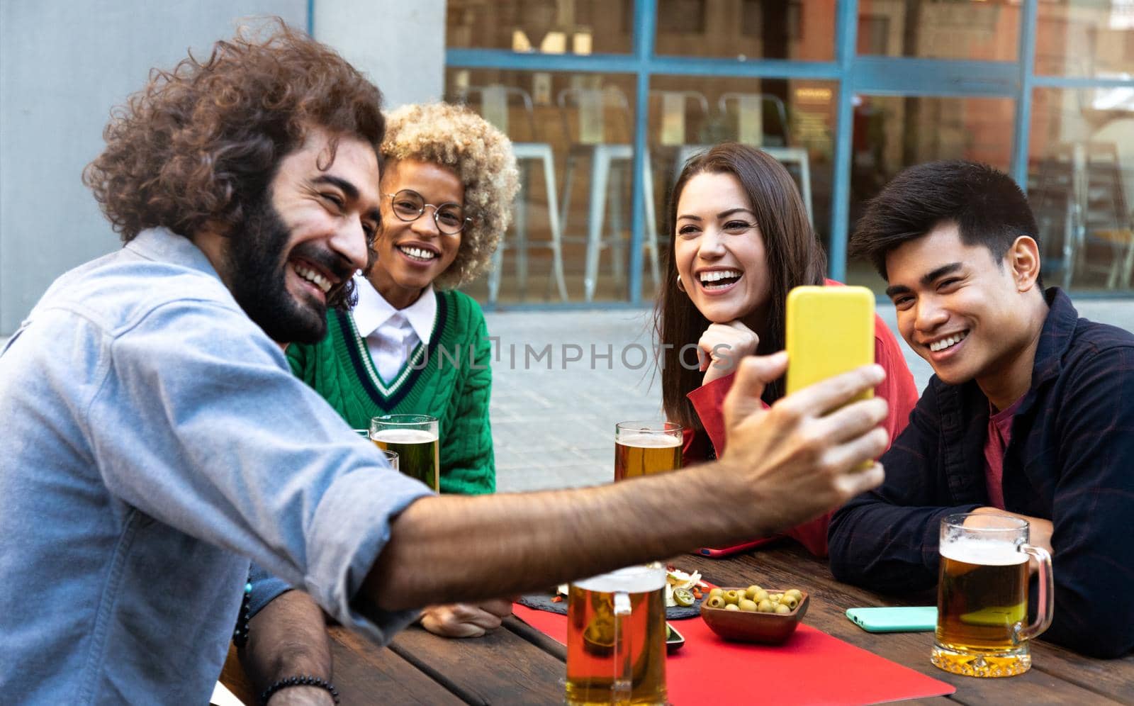Multiracial group of friends at a bar drinking beer look at cellphone together. Video call. Friendship and technology concept.