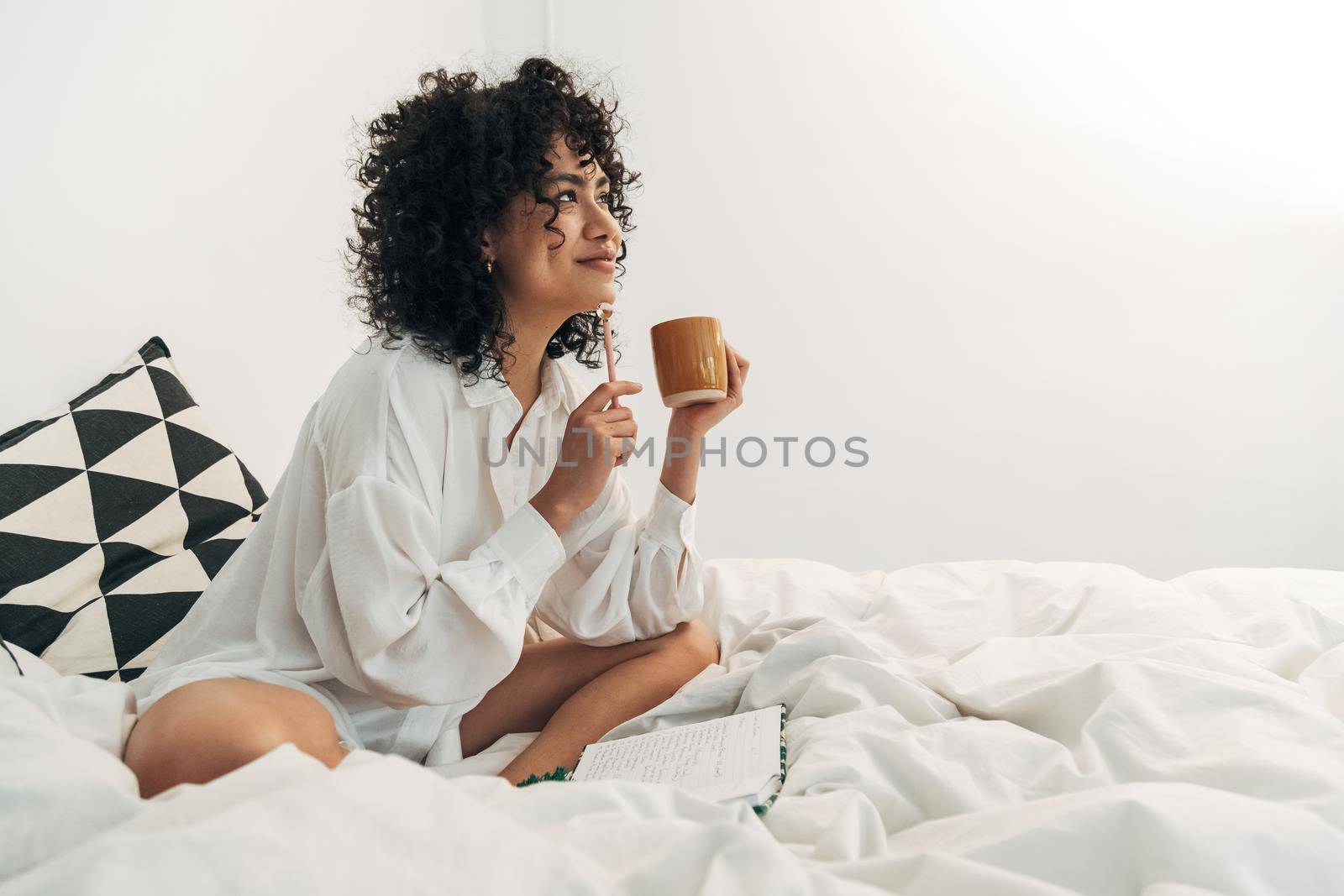 Young mixed race woman sitting on bed having coffee and writing on her journal. Copy space. by Hoverstock