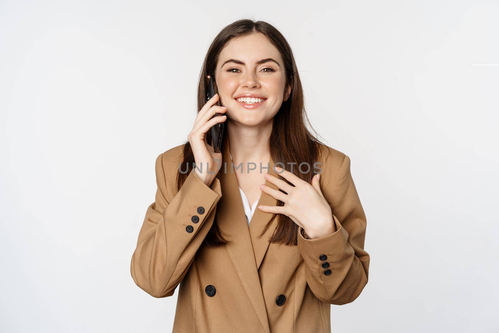 Businesswoman having mobile call, conversation on smartphone, talking with client, standing over white background in brown suit.