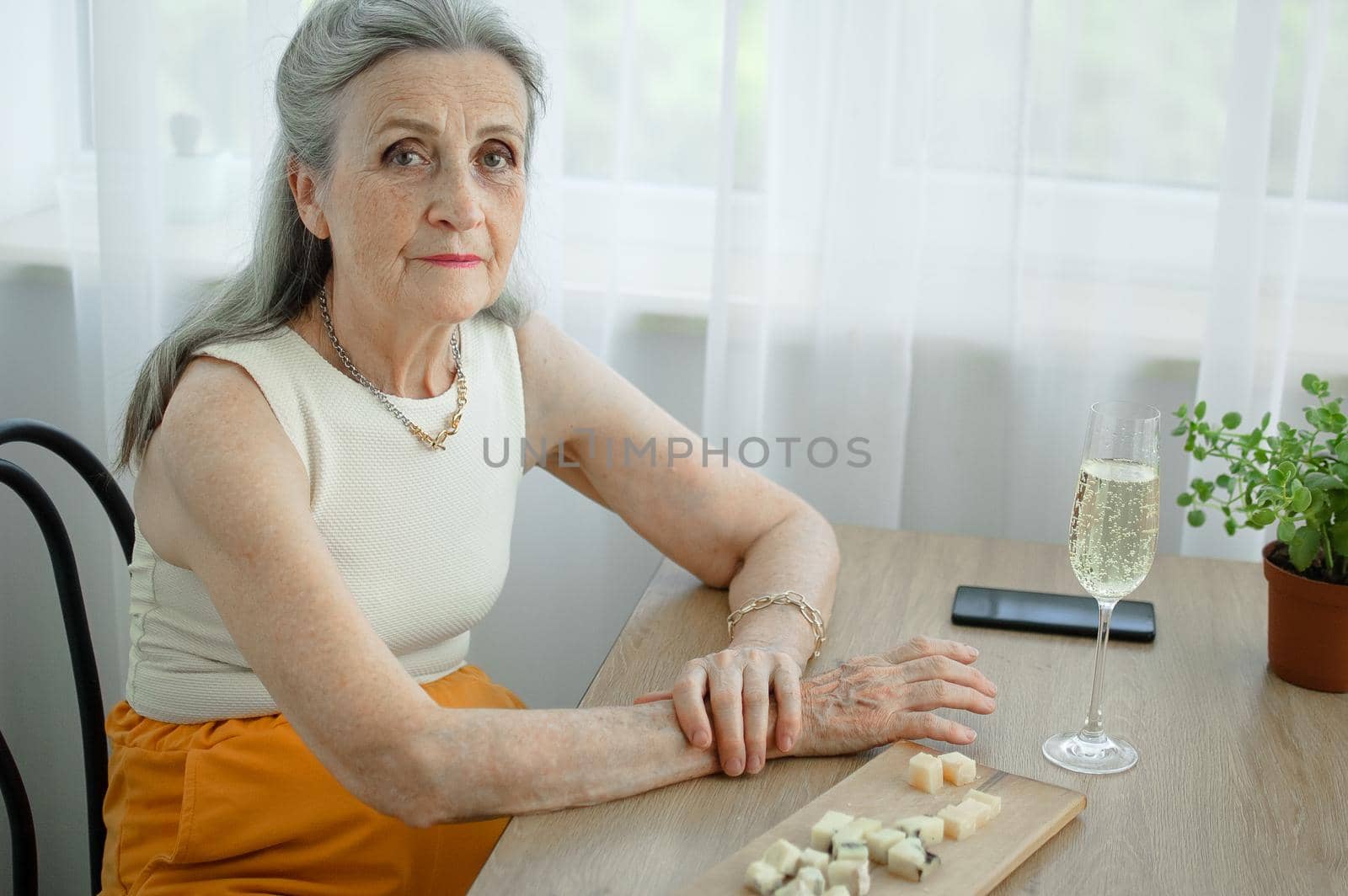 Beautiful old grandmother with grey hair and face with wrinkles sitting at the table at home on window background with glass of champagne, mother's day, happy retirement.