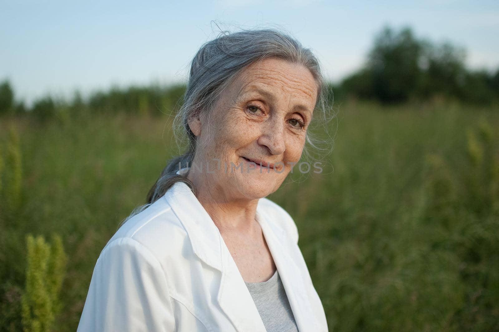 Portrait of senior woman with grey hair and face with wrinkles wearing white jacket and relaxing at park during sunny day, happy retirement, mothers day concepts