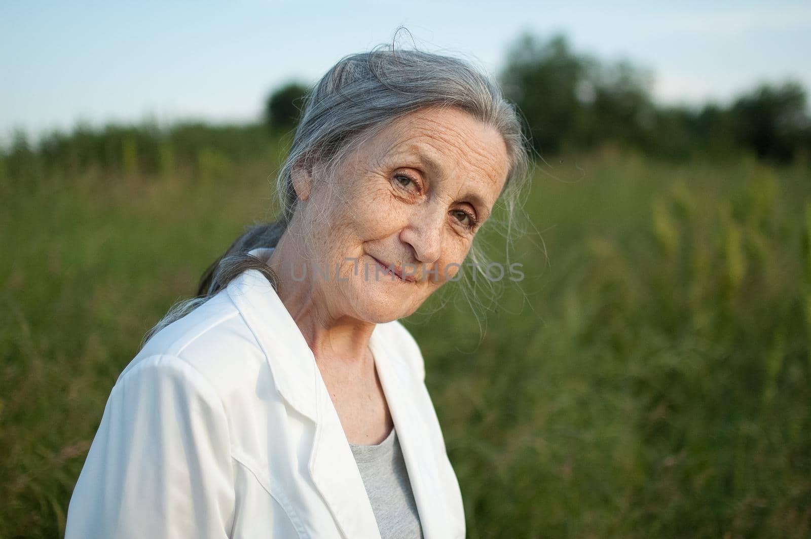 Portrait of senior woman with grey hair and face with wrinkles wearing white jacket and relaxing at park during sunny weather, mothers day by balinska_lv