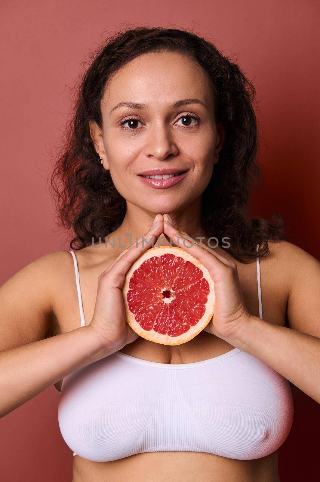 Beauty portrait of an attractive smiling middle aged beautiful woman with curly dark hair hair, wearing white underwear, isolated on bright coral background, holding half of fresh juicy red grapefruit by artgf