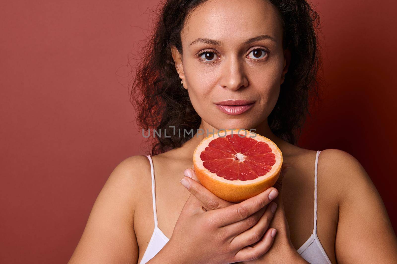 Close-up portrait of attractive woman holding half of fresh juicy red grapefruit, looking at camera, isolated over coral background. Healthcare and medicine, diet and healthy eating, lifestyle concept