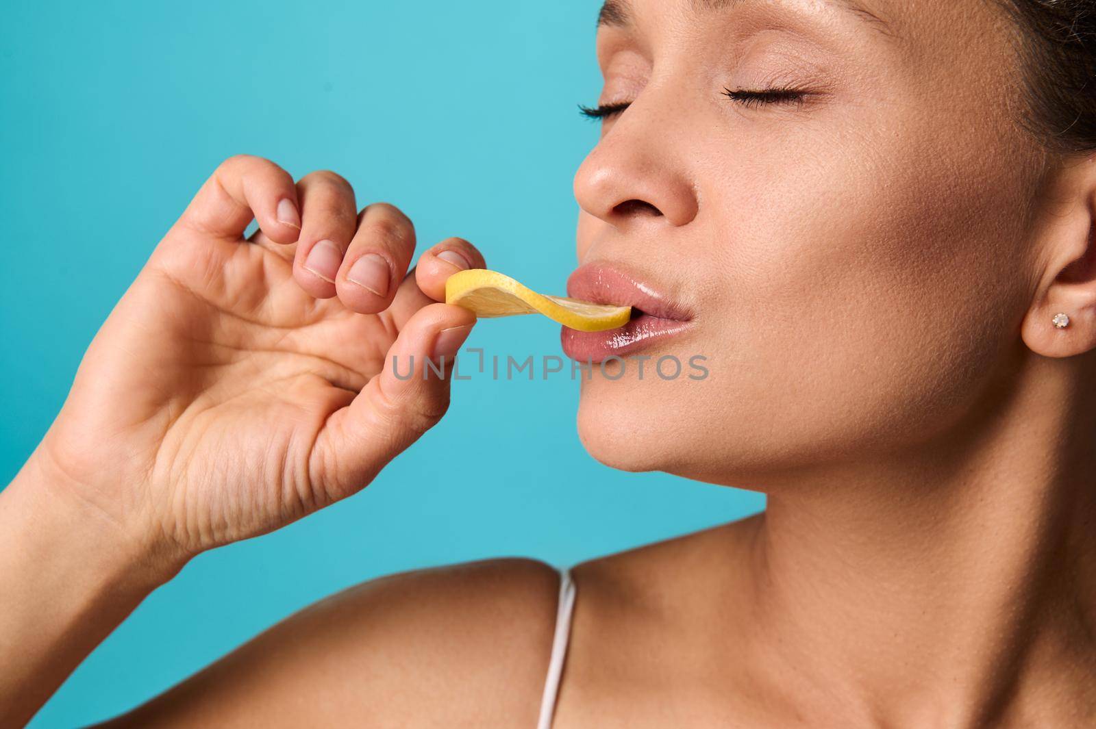 Headshot of a beautiful woman with healthy clean fresh glowing skin eating slice of lemon, posing with her eyes closed against bright blue colored background. Skin, body, health care concept. Close-up by artgf