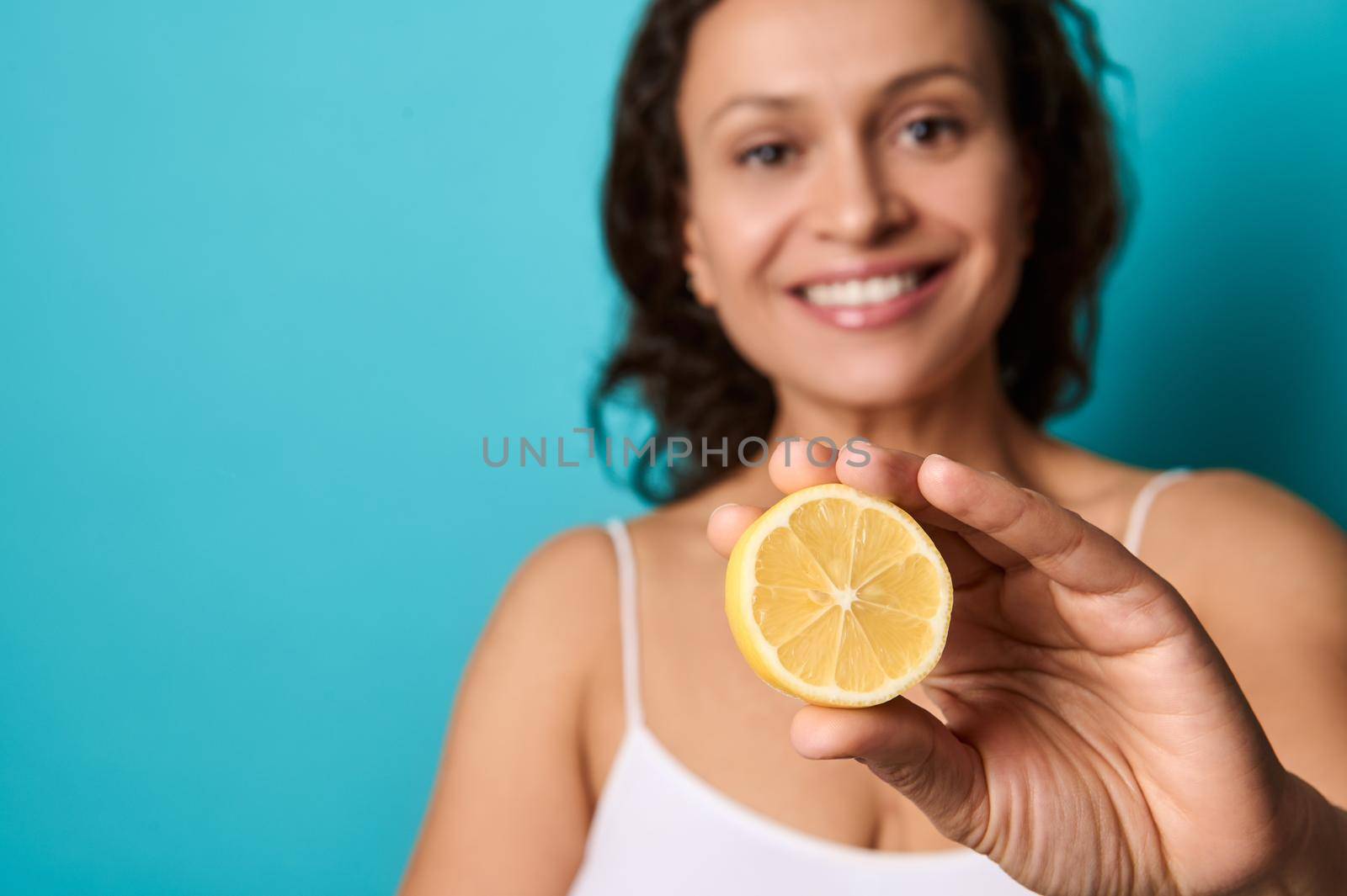 Beauty portrait of attractive cheerful smiling young woman with curly dark hair in white top isolated on bright blue background holding halves of lemon and covering her eyes with it. Copy ad space by artgf