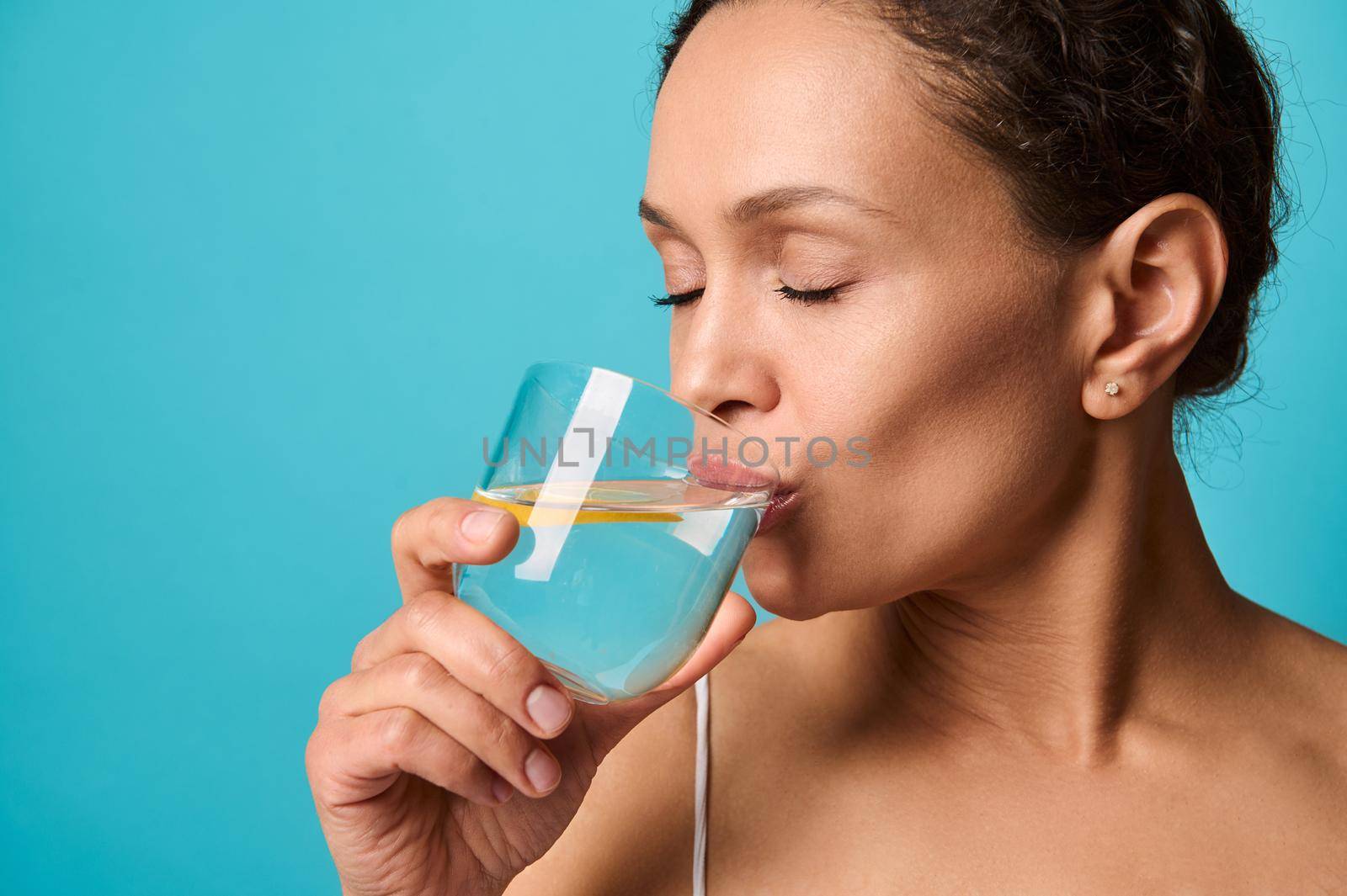 Headshot of a beautiful African woman with fresh healthy skin drinking lemon water from transparent glass, isolated over bright blue background with copy ad space. Healthy eating and lifestyle concept by artgf
