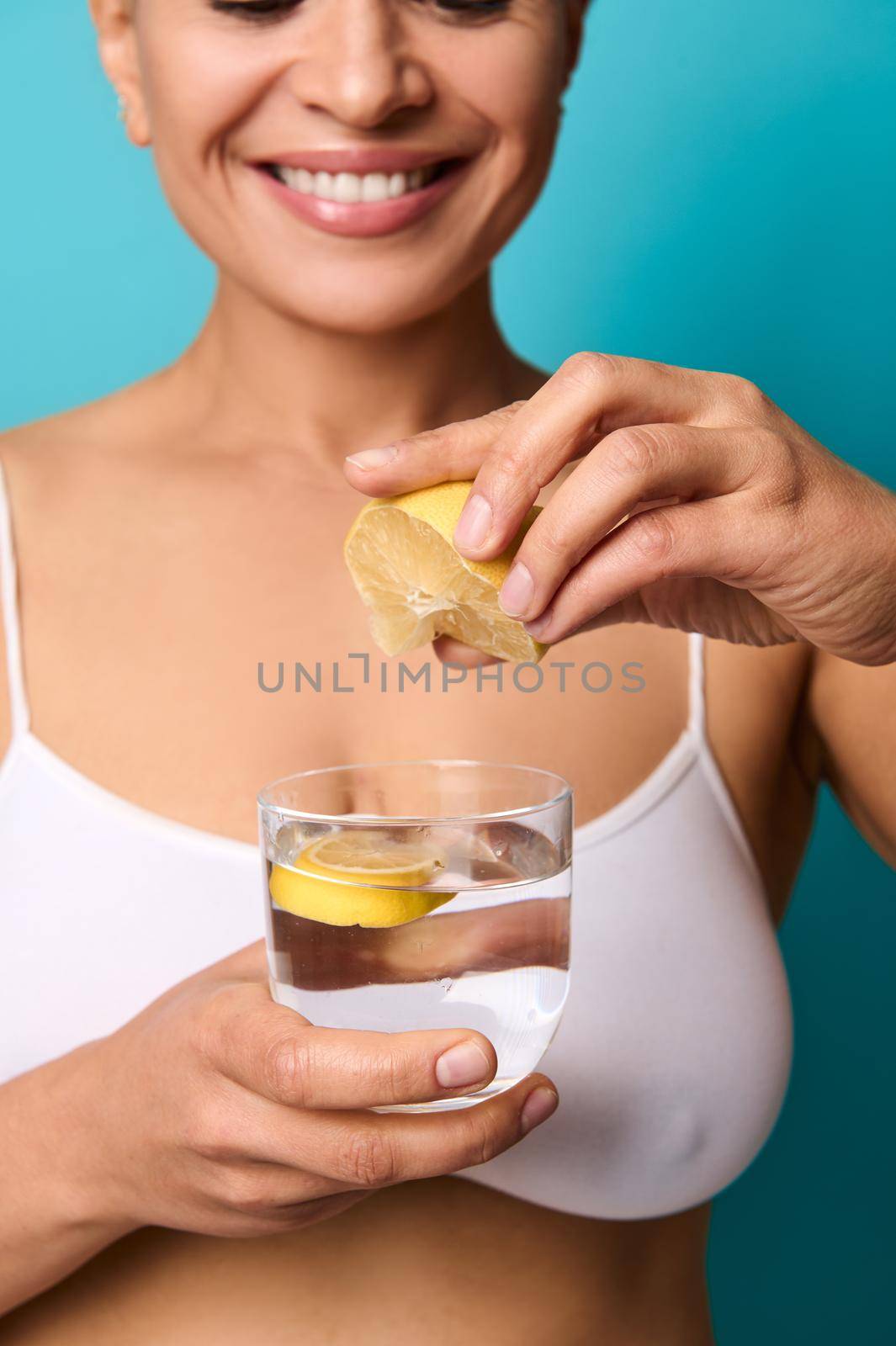 Soft focus in the hands of blurred pretty beautiful woman smiling toothy smile squeezing a fresh lemon juice into a glass with water, preparing lemon water, isolated on blue background with copy space by artgf