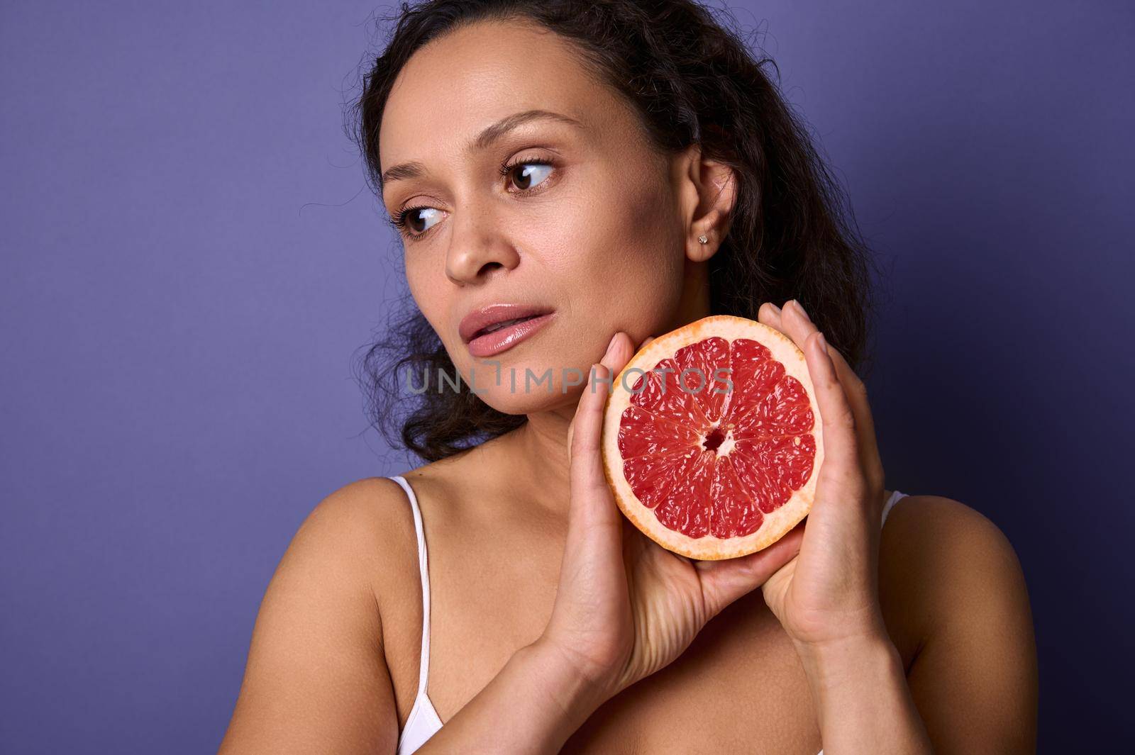 Close-up. Beautiful dark-haired woman with a half of red fresh juicy grapefruit near her face, looking away isolated over purple background with copy ad space. Health, body and skin care concept by artgf