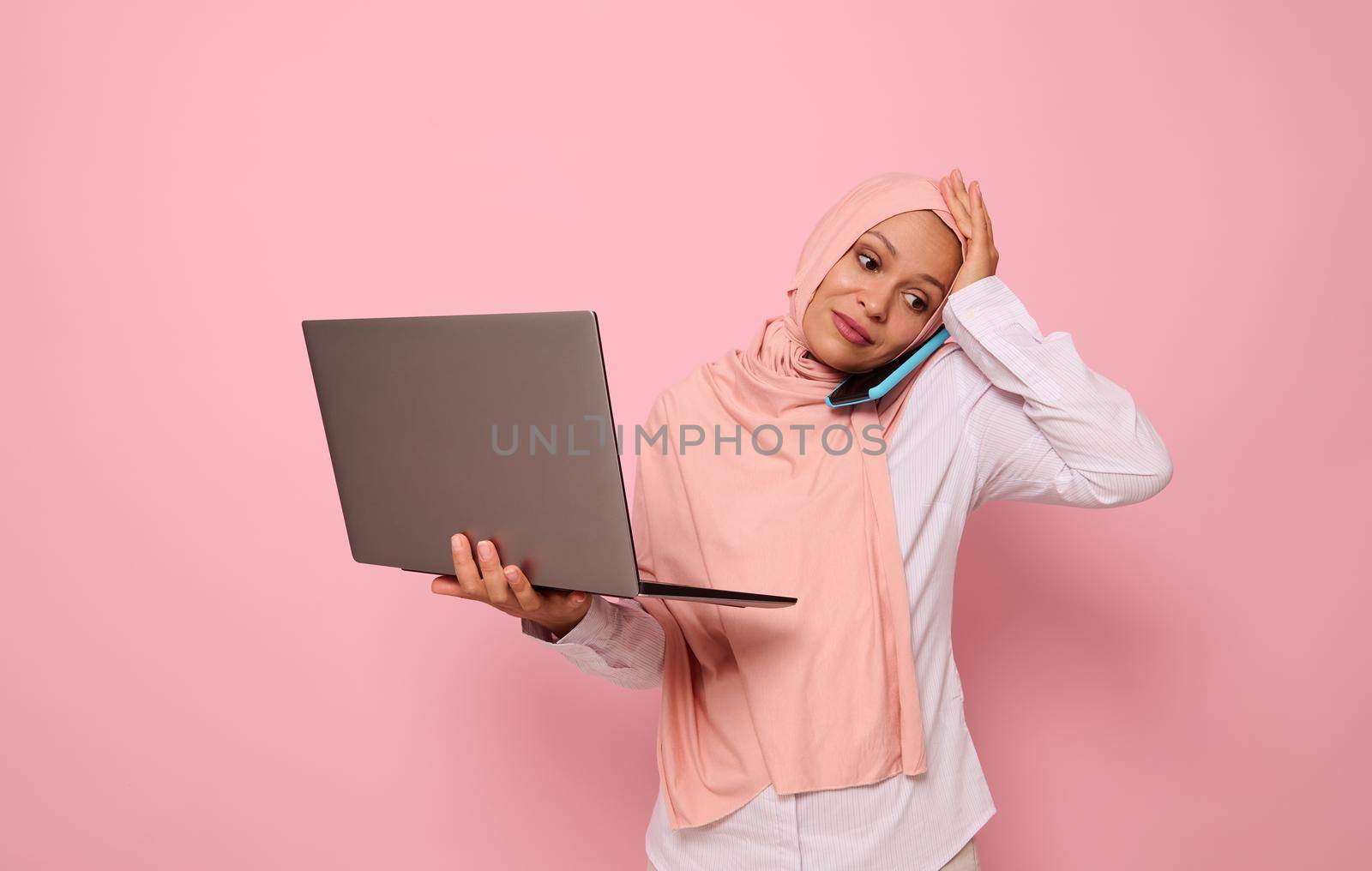 Portrait of puzzled Muslim Arab woman in pink hijab, with laptop, talking on the phone, holding her head because of multitasking, looking down with sad expression, on colored background copy space