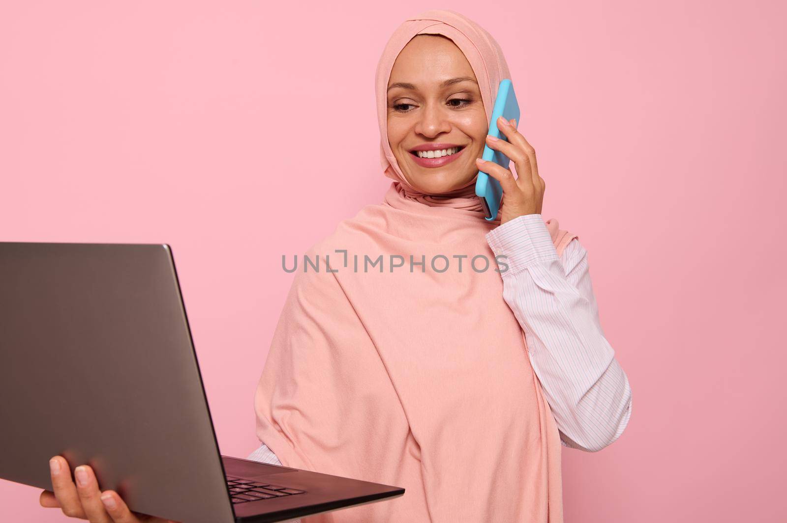 Close-up portrait of a beautiful Muslim Arab woman in pink hijab working on laptop, isolated on colored background with copy space. Successful programmer, IT female worker, start-up, business lady by artgf