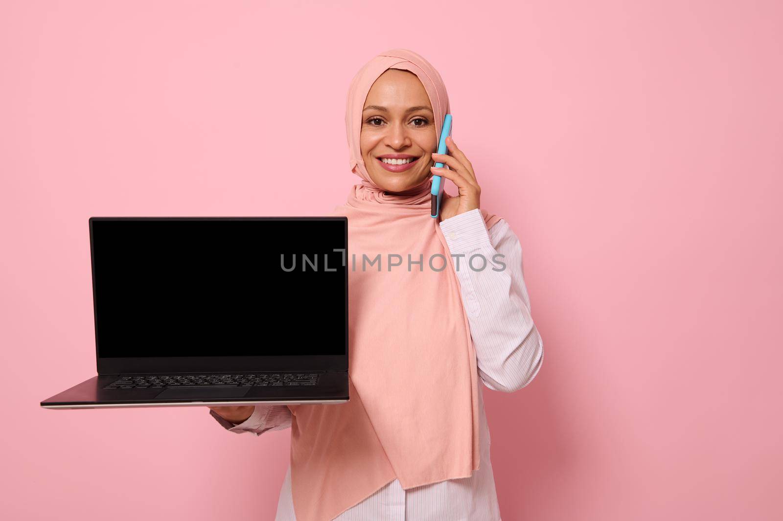 Pretty Arab Muslim woman in pink hijab talks on mobile phone, smiles with toothy smile and shows the empty blank black monitor screen with copy space to the camera. Isolated colored pastel background by artgf