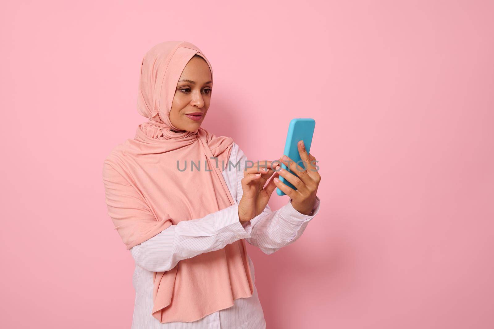 Isolated portrait of confident Arab Muslim mature pretty woman in strict religious outfit and covered head in pink hijab texting message on a mobile phone in her hands, colored background, copy space