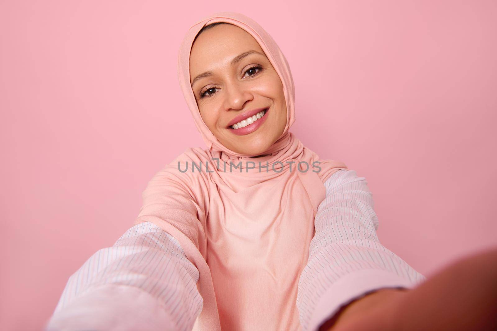 Close-up portrait of an attractive woman wearing traditional religious islamic outfit, pink hijab, holding smartphone in outstretched arms and smiles with toothy smile making a selfie, self-portrait by artgf