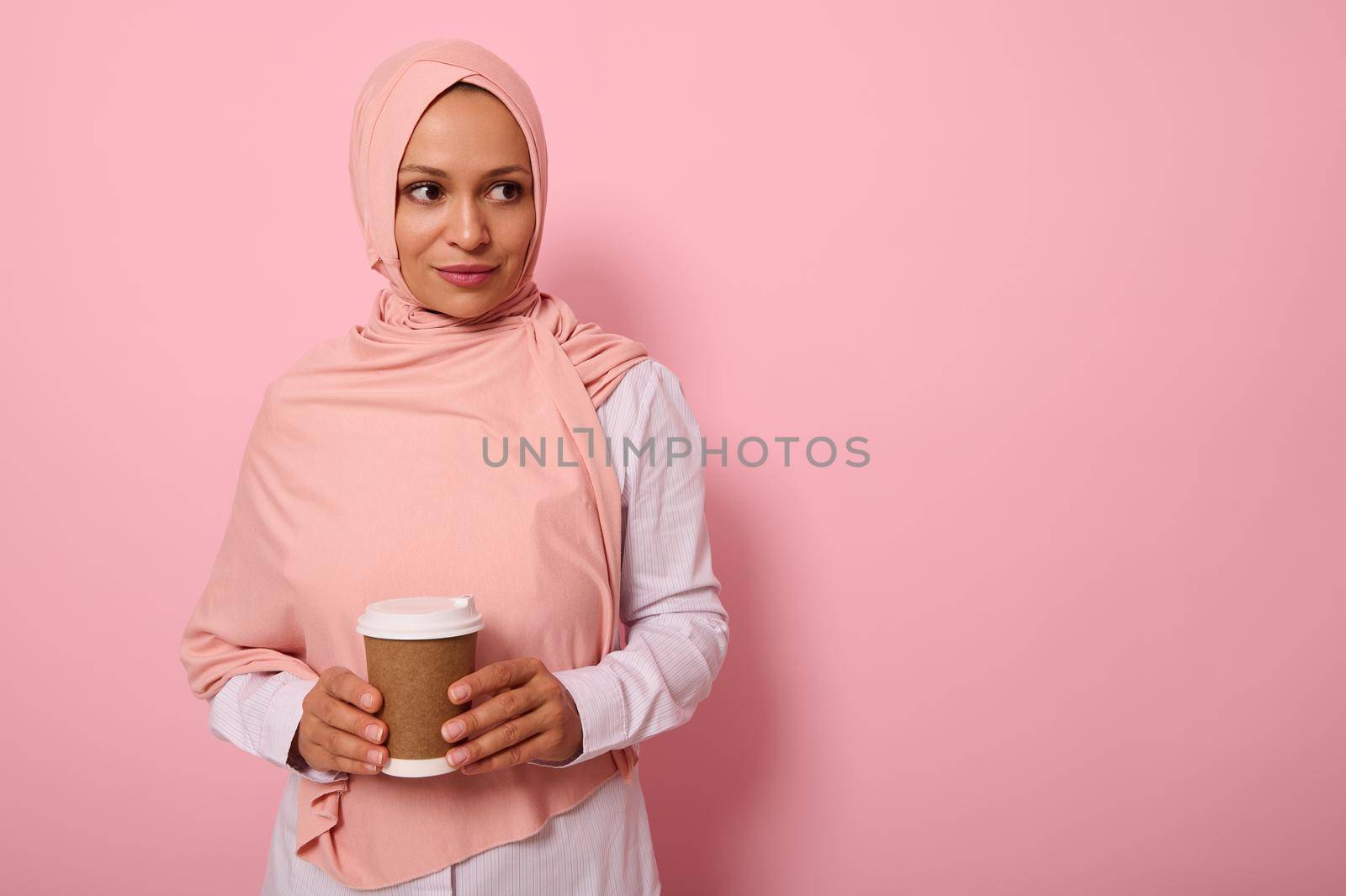 Arab Muslim beautiful woman wearing pink hijab and white shirt holding a recyclable disposable ecological paper mug in her hands, looking the side , isolated on colored background with copy space