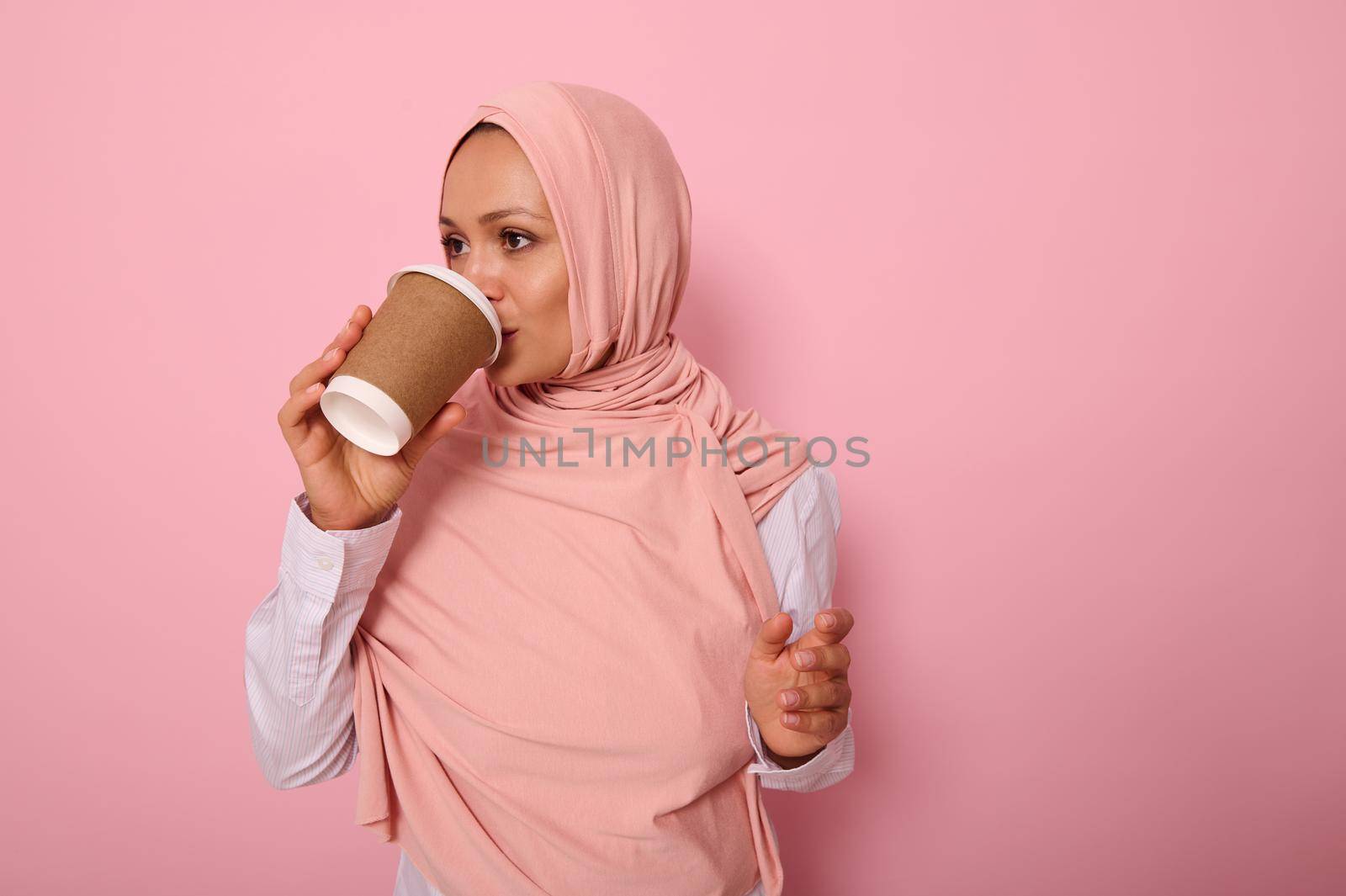 Beautiful Arabic muslim woman wearing a hijab drinking hot drink, tea or coffee from disposable cardboard takeaway cup, standing sideways, three quarters, against pink background with copy space