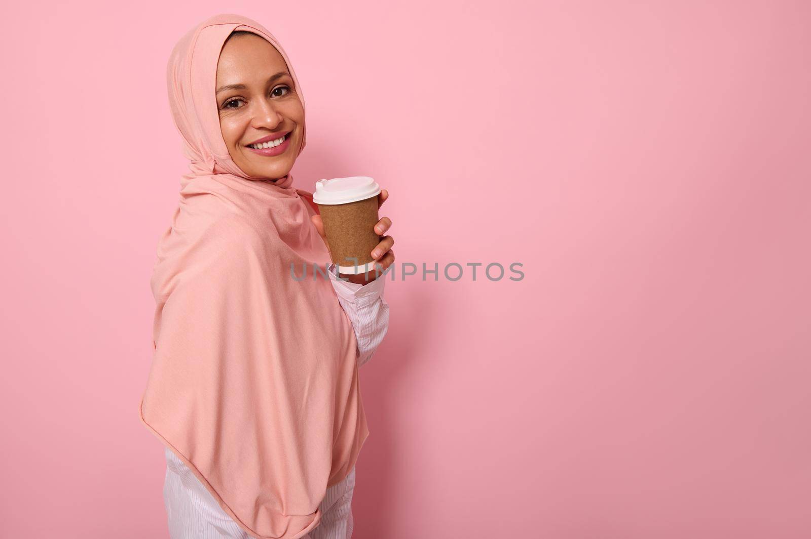 Arabic muslim cheerful woman with covered head in hijab stands three quarters against pink background with takeaway cup from disposable cardboard of hot drink, smiles looking at camera. Copy space
