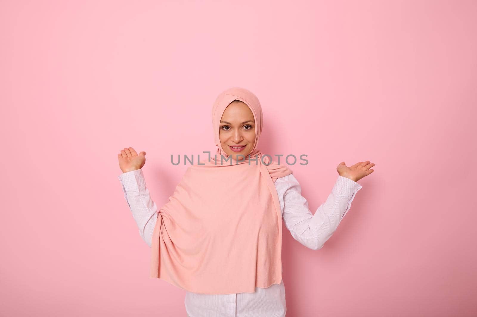 Young beautiful Muslim Arabic woman in pink hijab over isolated pink background holding a copy space imaginary on the palm to insert an advertising by artgf