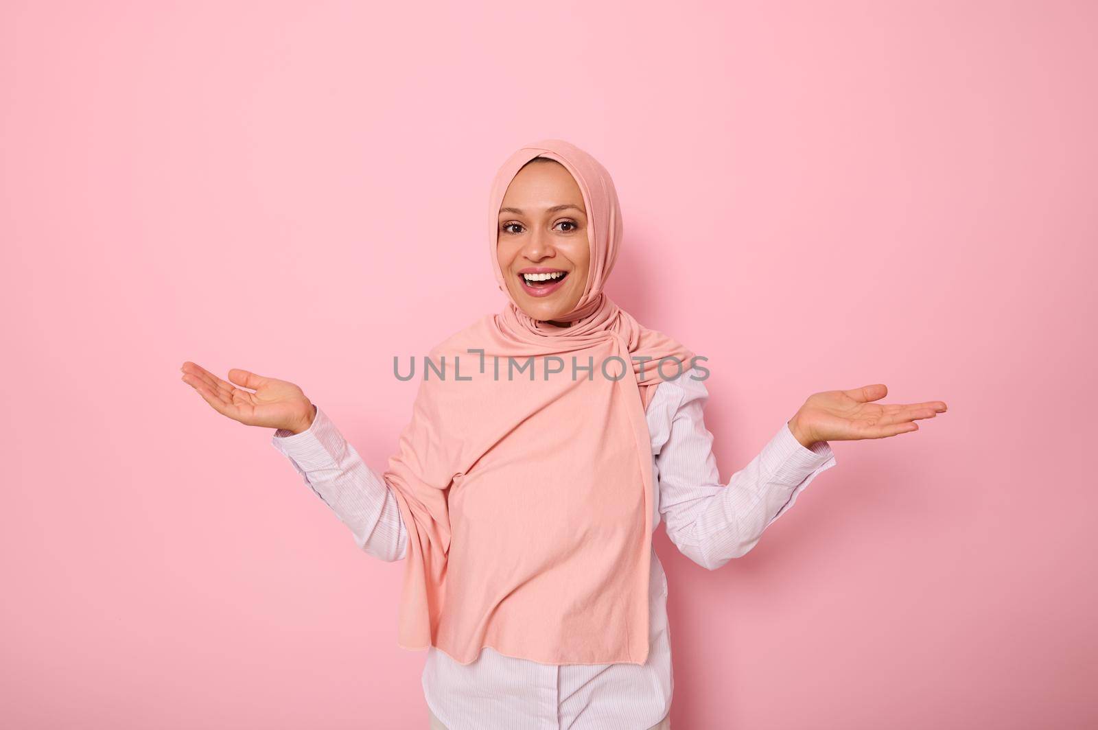 Pleasantly surprised gorgeous Arab muslim woman with covered head in pink hijab joyfully looks at camera smiling beautiful toothy smile while standing on a pink background with hands turned palm up by artgf