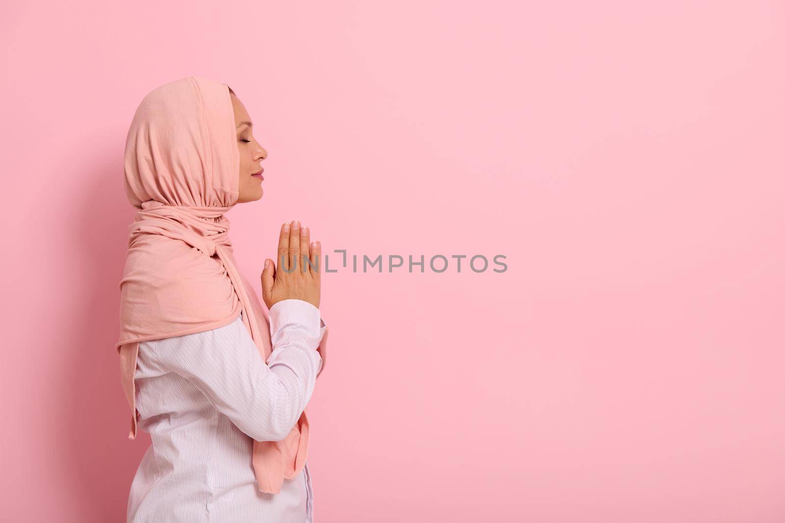 Side portrait of a serene Arab Muslim woman in pink hijab and strict outfit with palms folded together at chest level praying, performing namaz, isolated on colored background with space for text