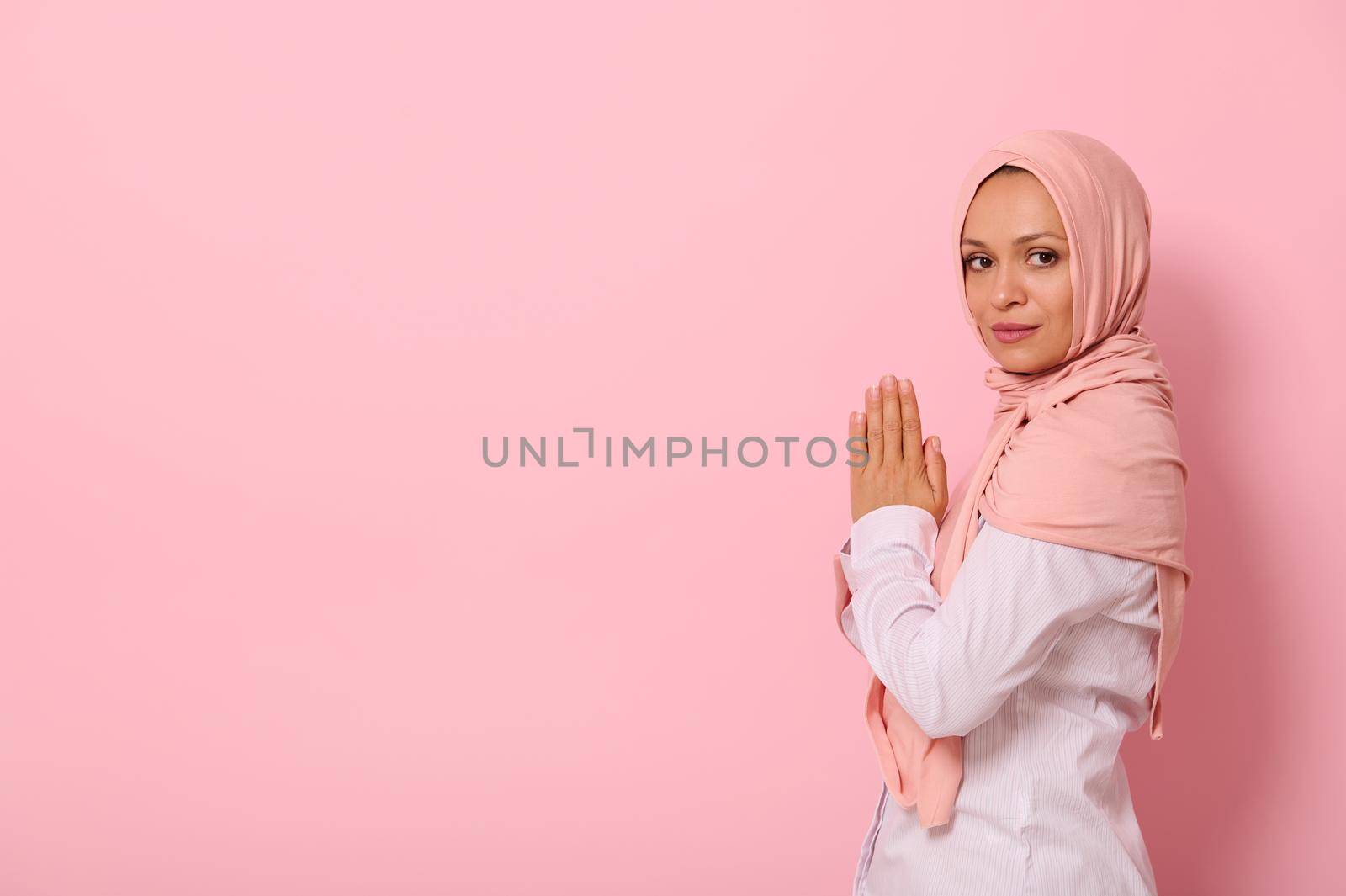 Muslim Arab woman in pink hijab and strict outfit with palms folded together at chest level praying, performing namaz, looking confidently at camera, isolated on colored background with space for text by artgf