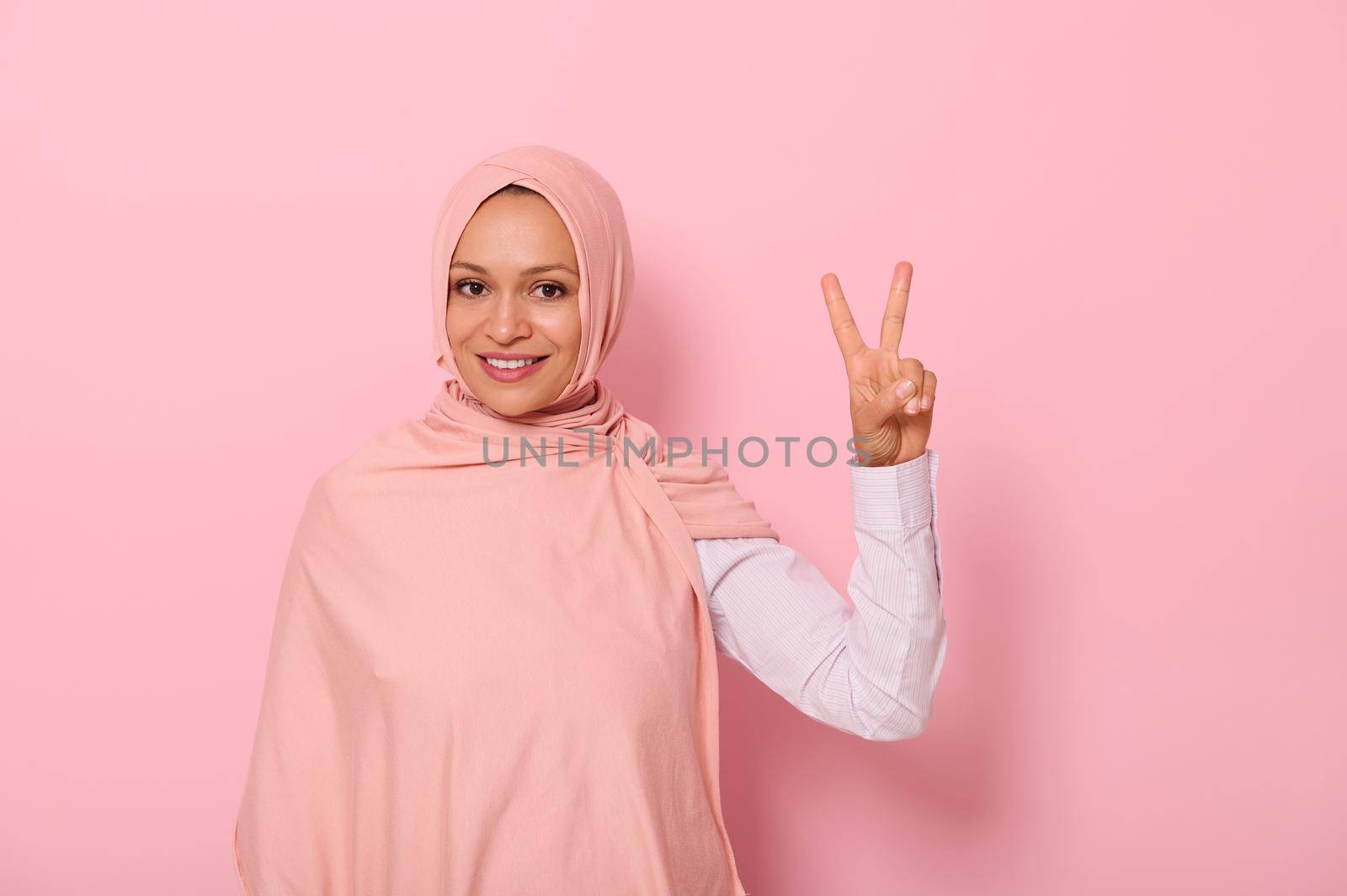 Attractive friendly Arab Muslim woman in colored hijab showing finger peace sign, smiling with toothy smile looking at camera, isolated on pink background with copy space by artgf