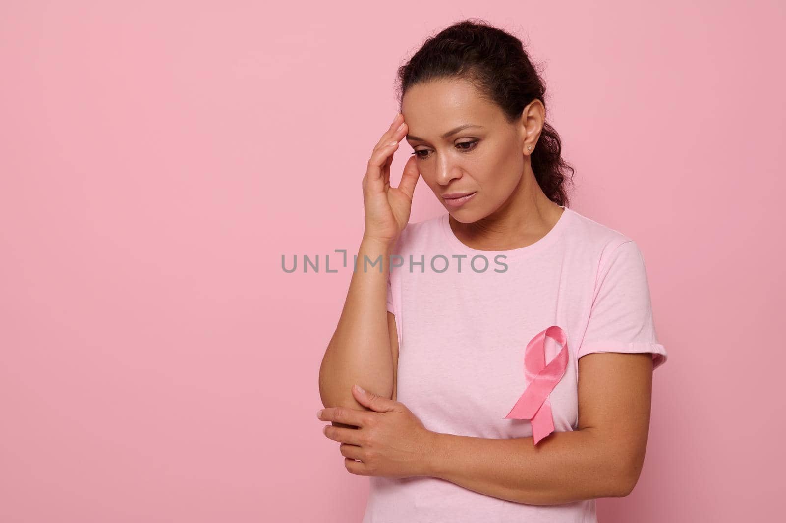 Beautiful thoughtful young woman in pink t-shirt with pink ribbon looking down on pink background, hands on temple. International World Breast Cancer Awareness Day, Educational and medical concept.