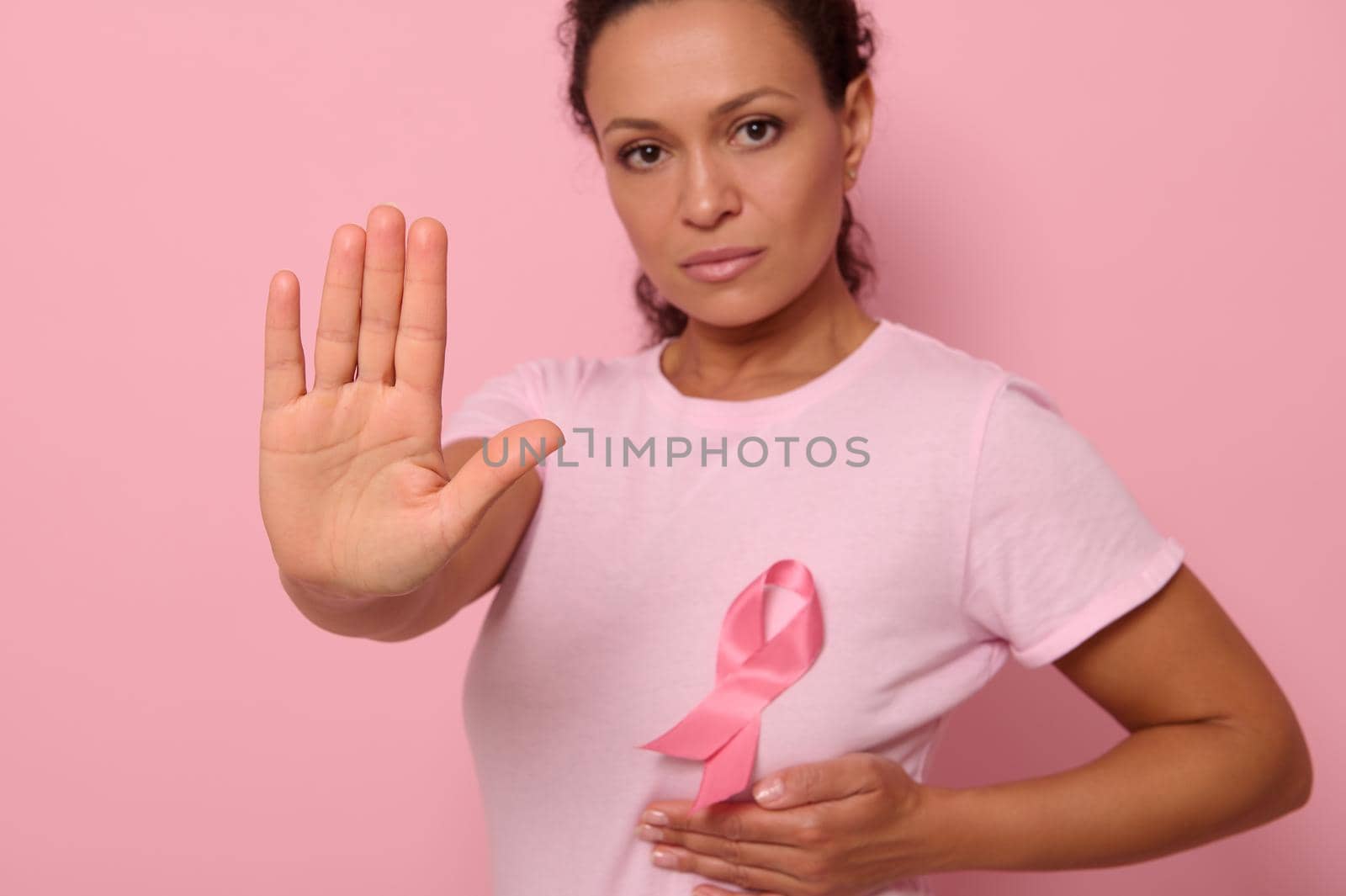 Confident portrait of blurred woman wearing pink t-shirt and cancer awareness ribbon. Focus on woman's hand gesturing STOP , isolated over pink background with copy space by artgf