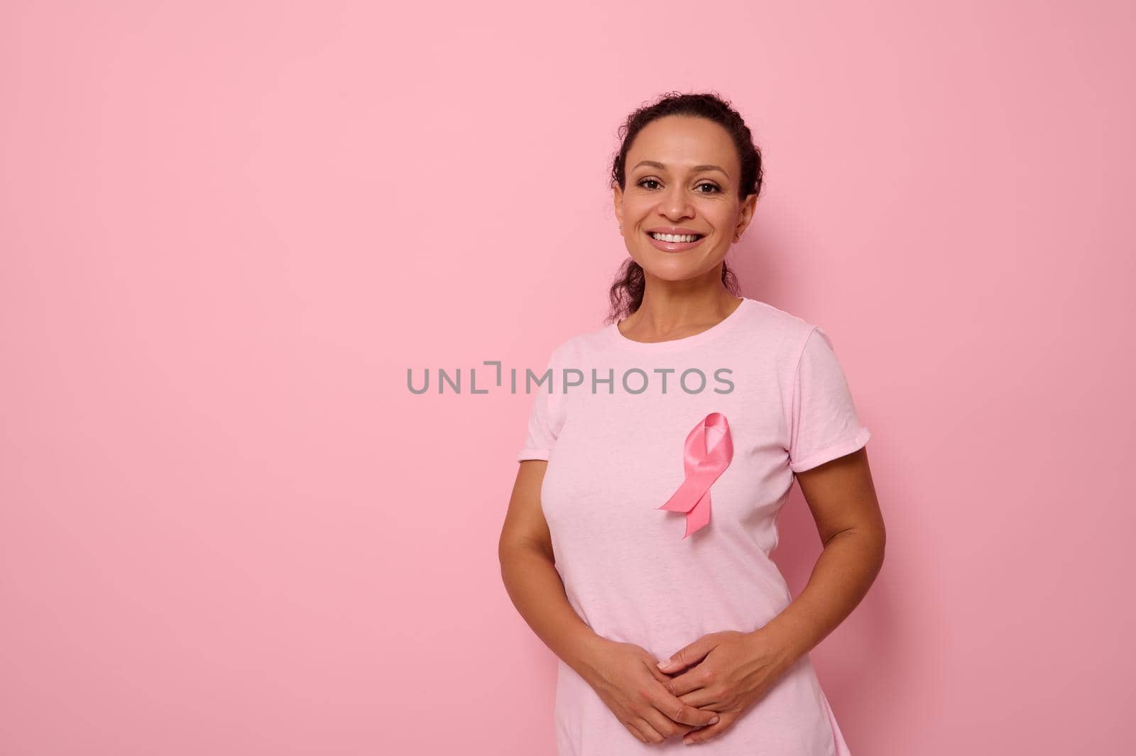 Smiling mixed race woman in pink T-shirt with pink satin ribbon symbolizing International Breast Cancer Day, expressing solidarity and support for breast cancer patients and survivors. 1 st october by artgf