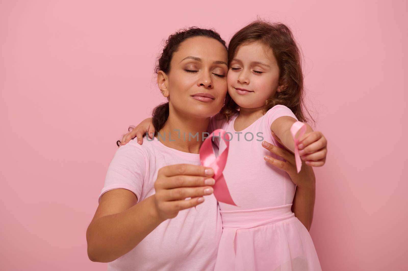 Adorable mixed-race woman mother hugs a little girl in pink outfits and displays a pink satin ribbon to camera, symbol of Breast Cancer Day in October Month. Support for cancer patients and survivors by artgf