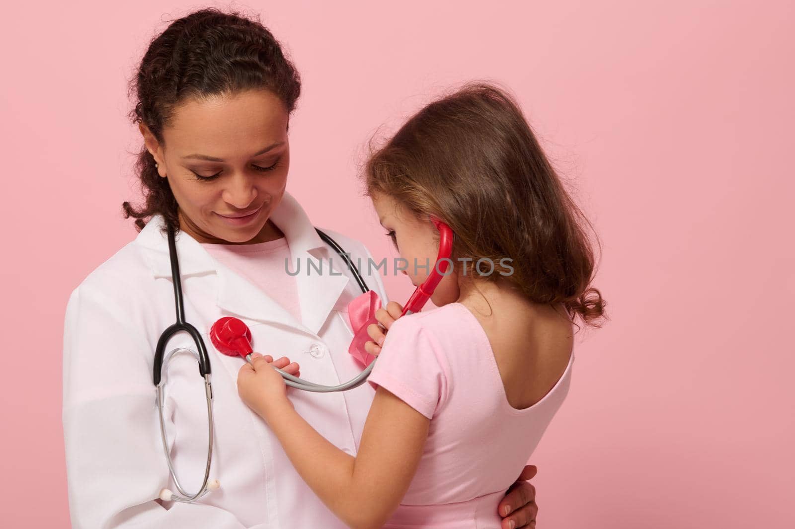 Beautiful 4 y.o. child girl playing doctor, using stethoscope, listening to chest and lungs breathing of doctor with pink ribbon on chest. Concept of educational program for supporting cancer patients