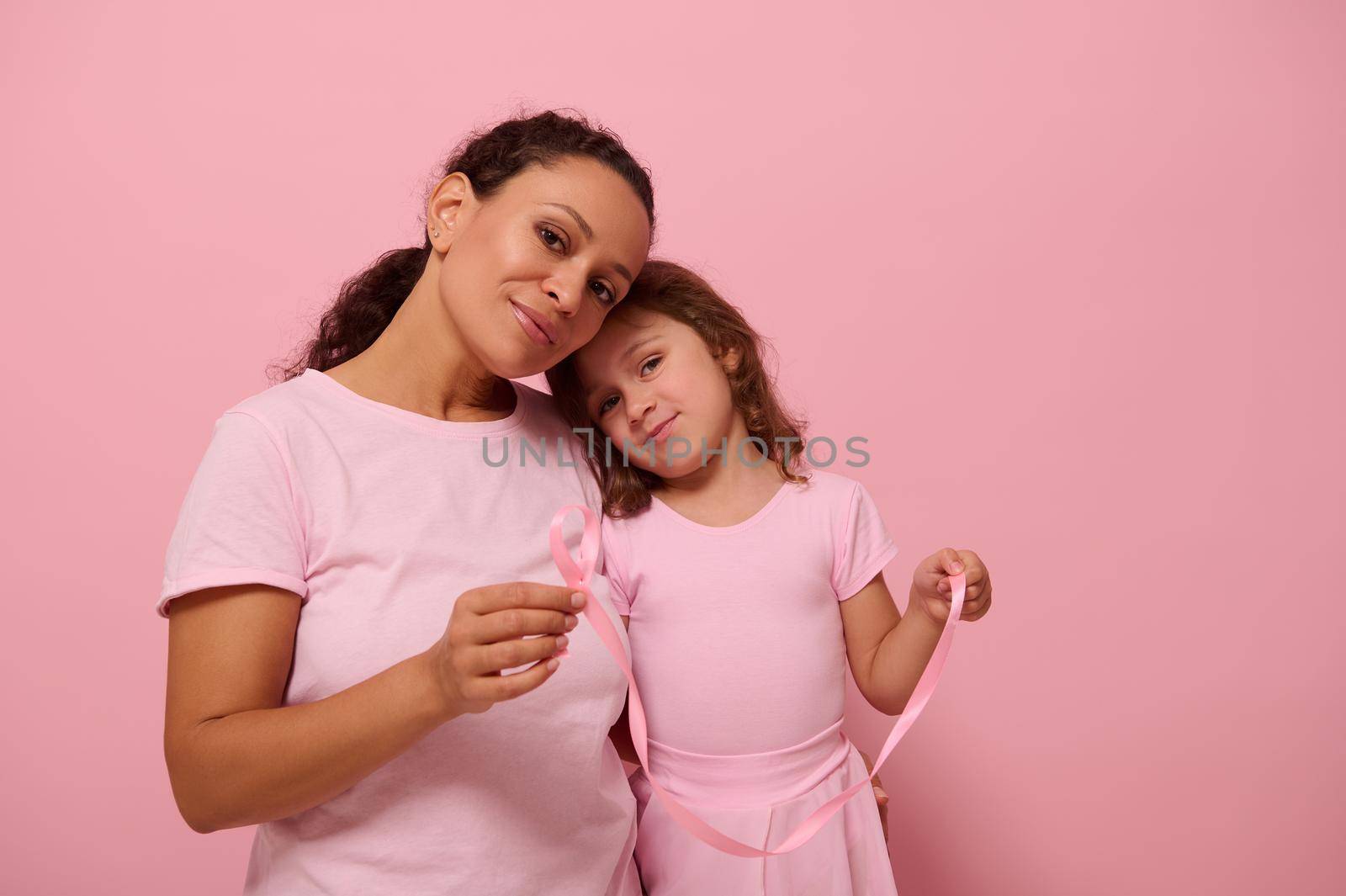 Adorable baby girl , leans on her mother shoulder,both dressed in pink color outfit, hold pink satin ribbons, cute smile looking at camera . World Breast Cancer Awareness Day. Fight Cancer. Copy space by artgf