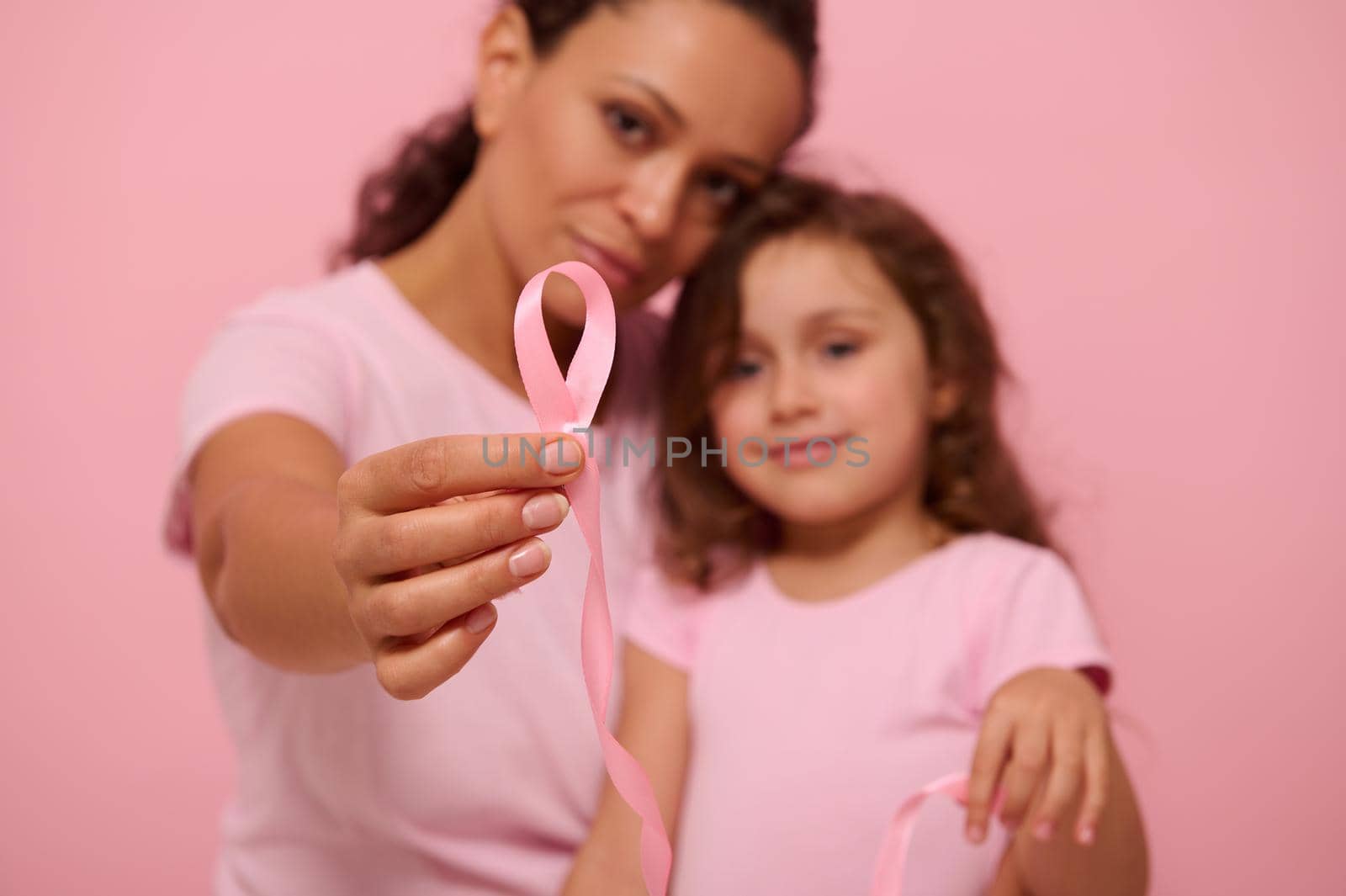 Focus on a long pink satin ribbon with one endless end in the hands of blurred two generations of pretty women, mom and daughter, expressing support and solidarity for cancer patients and survivors by artgf