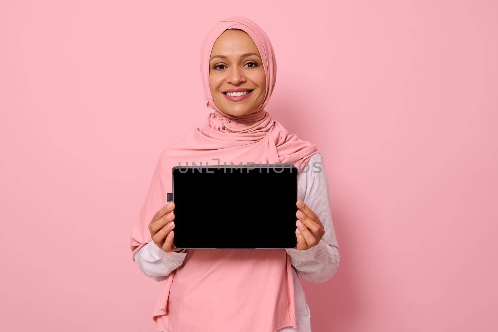 Arab Muslim beautiful woman wearing pink hijab, smiling toothy smile looking at camera, standing against pink background with copy space and showing the empty blank screen of a digital tablet by artgf