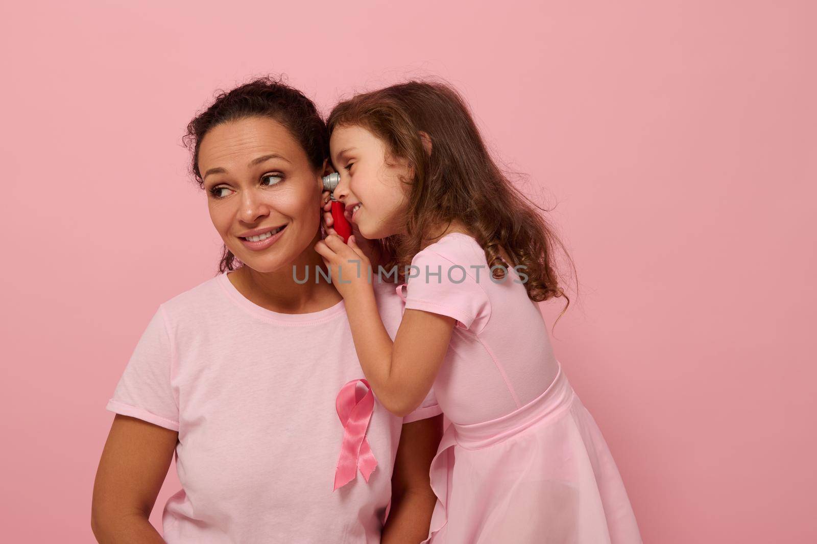 Little girl playing doctor with her mother. Beautiful African American woman with baby girl in pink with pink ribbon, symbol of International Breast Cancer Awareness Day. Medical Educational concept by artgf