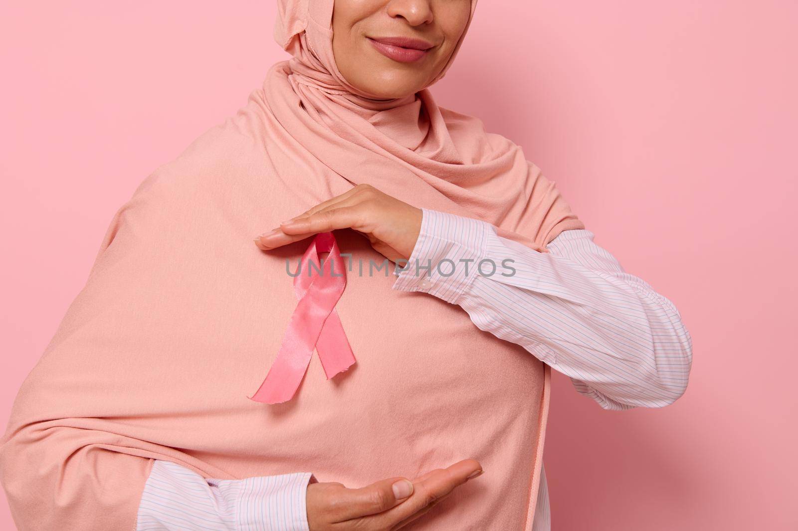 Cropped view. Muslim woman wearing hijab holding pink satin ribbon at chest level to show her support for cancer patients and survivors. World Breast Cancer Awareness Day. Pink background, copy space by artgf