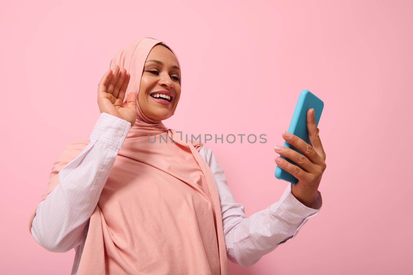 Muslim cute woman of Middle Eastern or Arab ethnicity with covered head in pink hijab having a video conference on mobile phone, greeting her interlocutor during video call, pink background copy space by artgf
