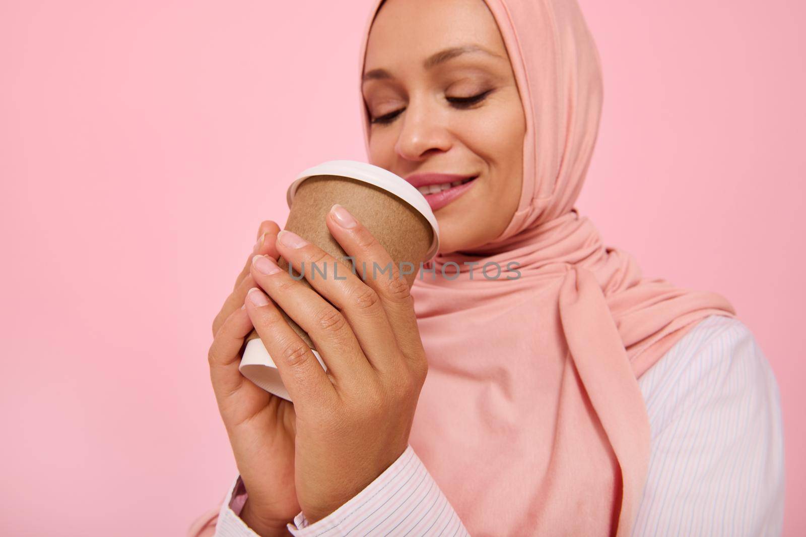 Arabic Muslim woman with covered head in pink hijab drinking hot drink, tea or coffee from disposable cardboard takeaway cup, standing three quarters against colored background, copy space. Close-up by artgf