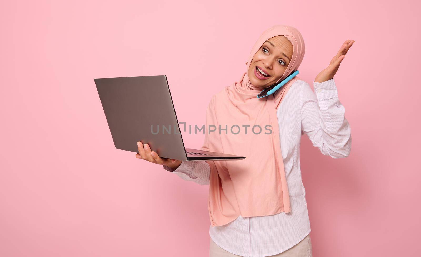Busy and puzzled Muslim Arab gorgeous pretty woman in hijab, dressed business smart casual holds a laptop and talks on mobile phone , gestures with hands. Isolated over pink background with copy space