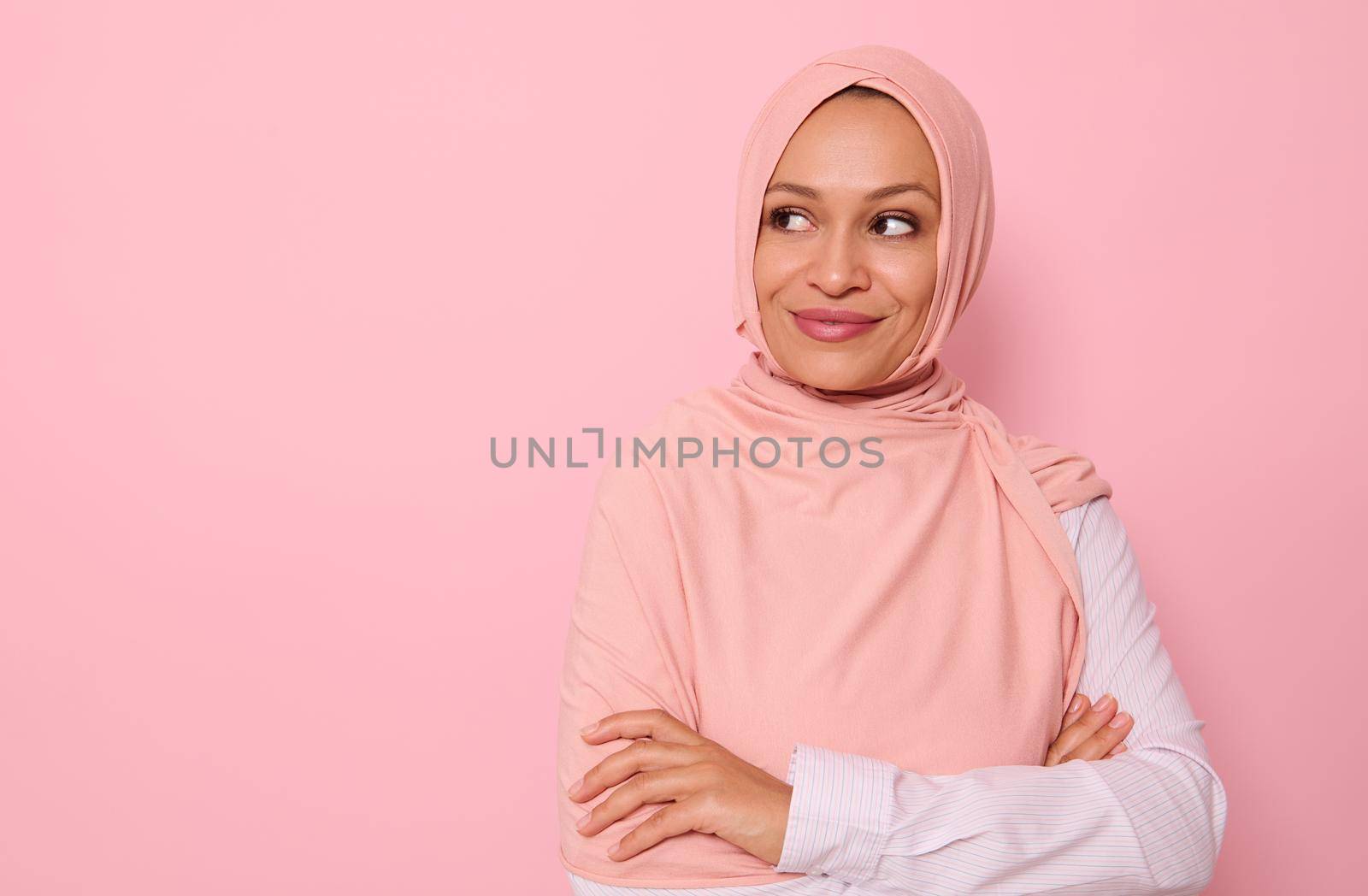 Close-up portrait of a middle aged Arab Muslim pretty gorgeous woman with covered head in hijab looking mysteriously at the side on a pink background with copy space and smiling thoughtfully by artgf