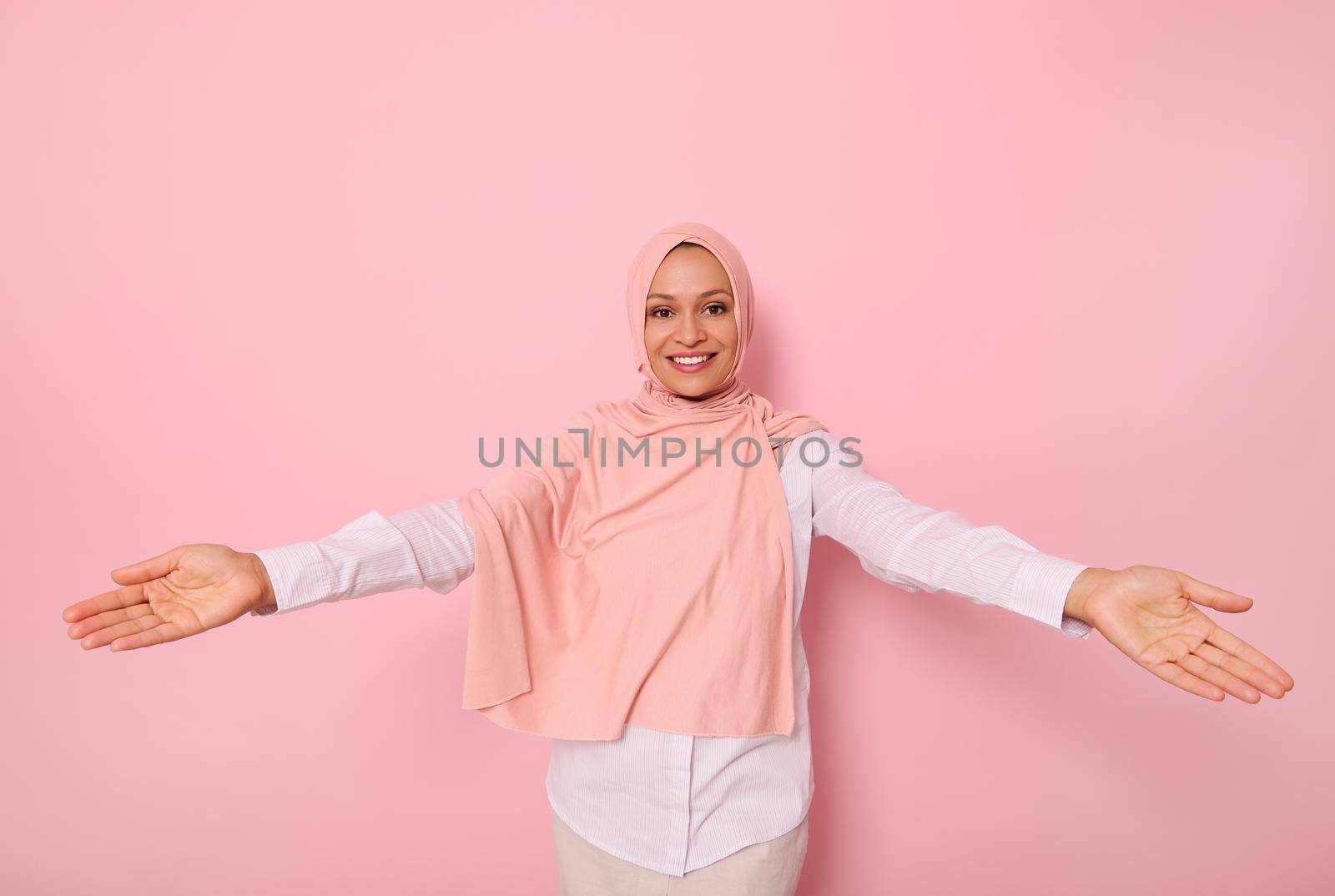 Friendly, cute smiling with a charming beautiful toothy smile young Muslim Arab woman in pink hijab looking at camera with outstretched arms showing hospitality, on colored background with copy space