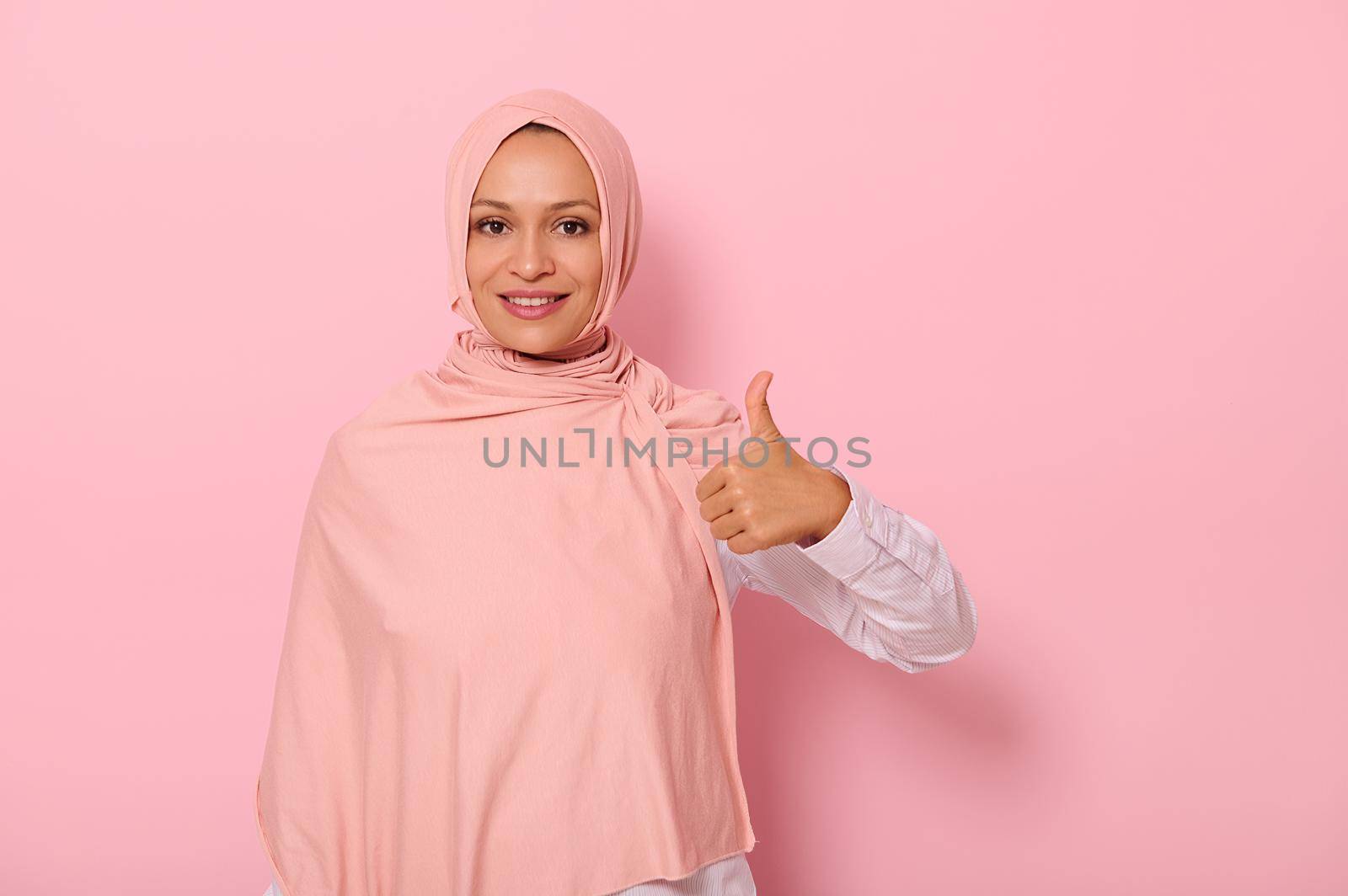 Isolated portrait on pink background with copy space of an adorable and charming Arabic woman in hijab showing a thumb up and smiling with beautiful toothy smile looking at the camera