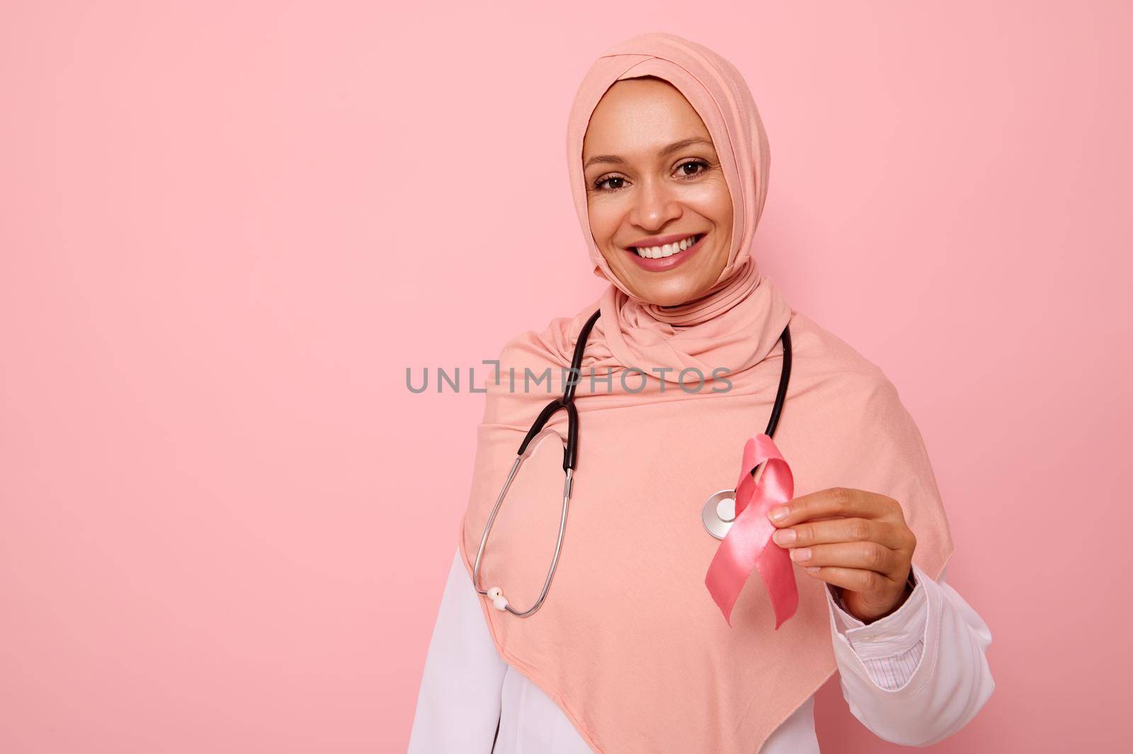 Gorgeous Arab Muslim female doctor in hijab with beautiful toothy smile showing Pink satin Ribbon, isolated on colored background, copy space. World Day of fight Breast Cancer, 1 st October concept